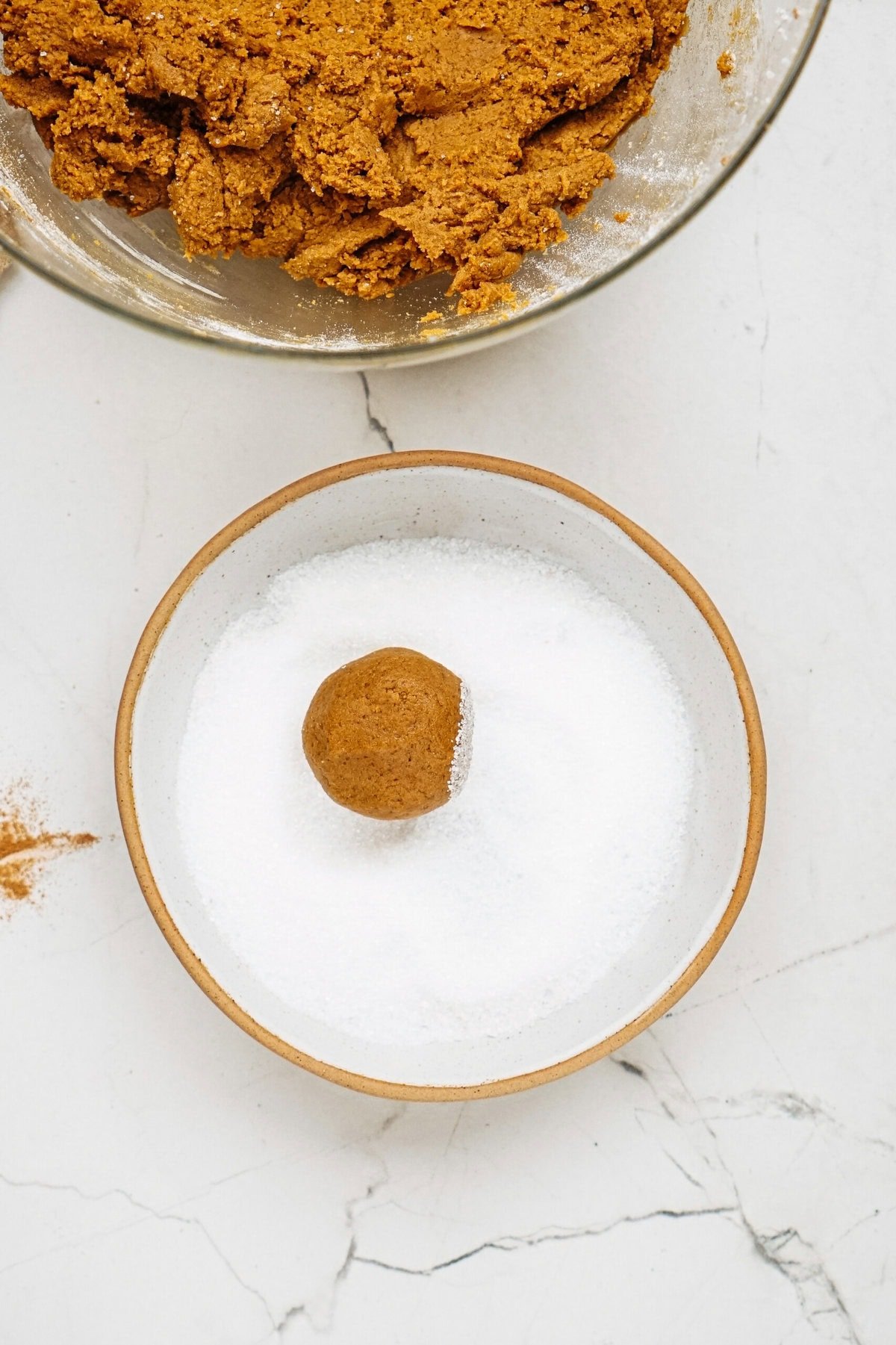 A ball of dough is placed in a bowl of white sugar. Another bowl with more dough is partially visible above on a marble surface.