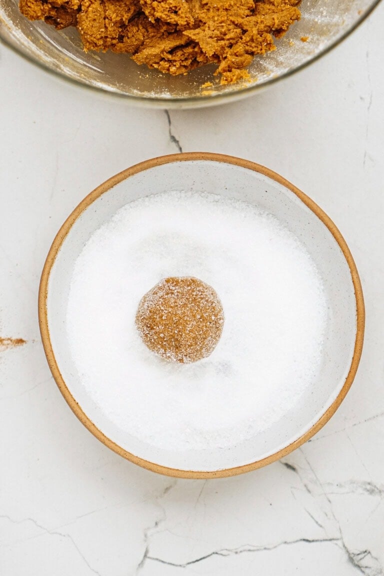 A cookie dough ball is partially coated in white sugar, placed in a ceramic bowl on a marble surface. Another bowl with dough is visible at the top.