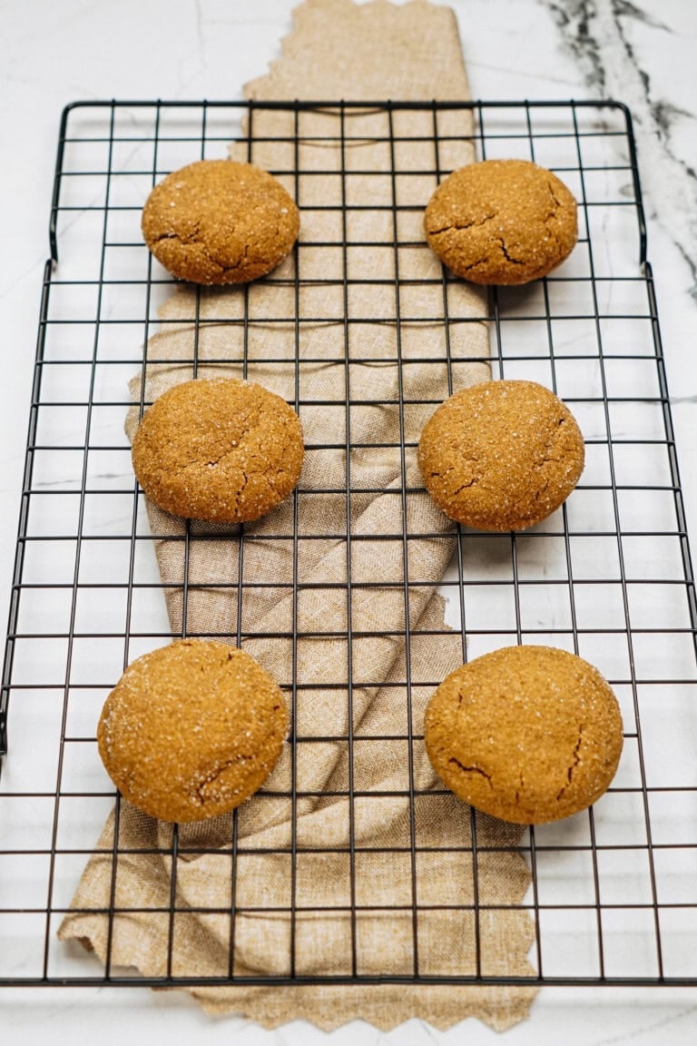 Six brown cookies on a cooling rack over a textured beige cloth.