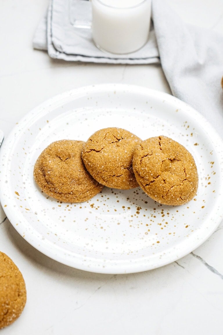 Three ginger cookies on a white speckled plate, with a napkin and a glass of milk in the background.