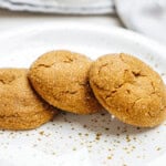 Three sugar-coated ginger cookies on a white speckled plate, with a blurred background featuring a glass of milk.