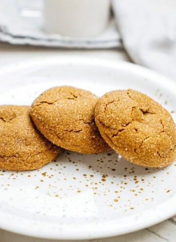 Three sugar-coated ginger cookies on a white speckled plate, with a blurred background featuring a glass of milk.