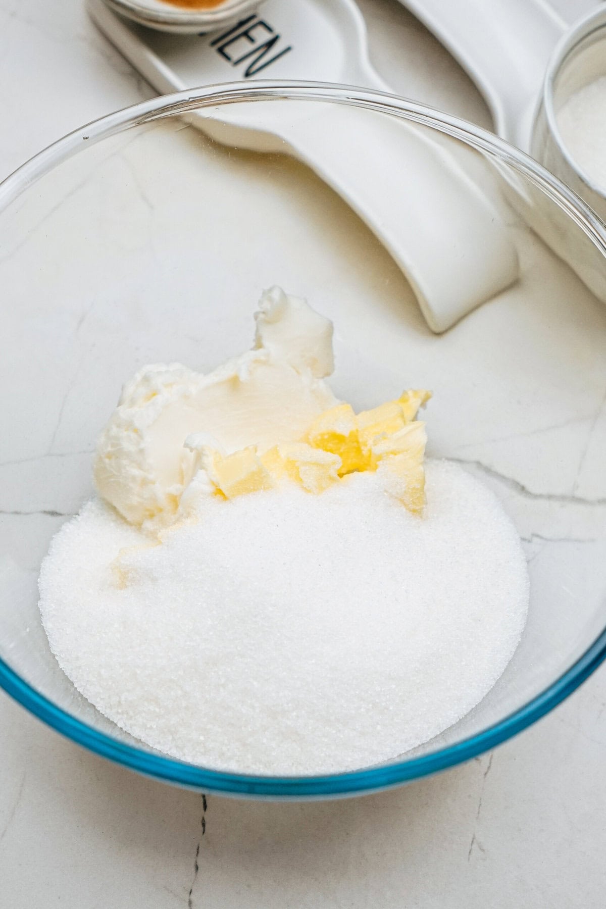 A glass bowl contains sugar, butter, and cream cheese on a marble surface. Measuring cups are visible in the background.