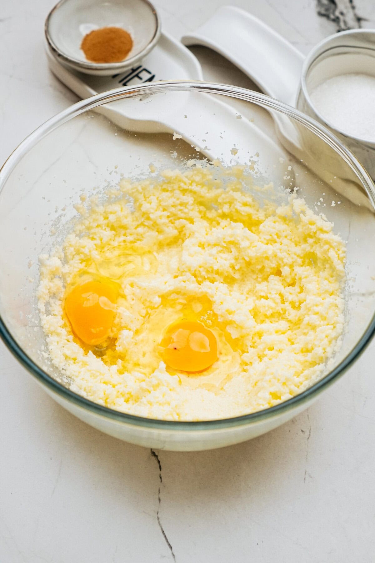 A glass bowl with creamed butter and sugar mixture, topped with two cracked eggs. Nearby are small bowls containing cinnamon and sugar on a marble countertop.