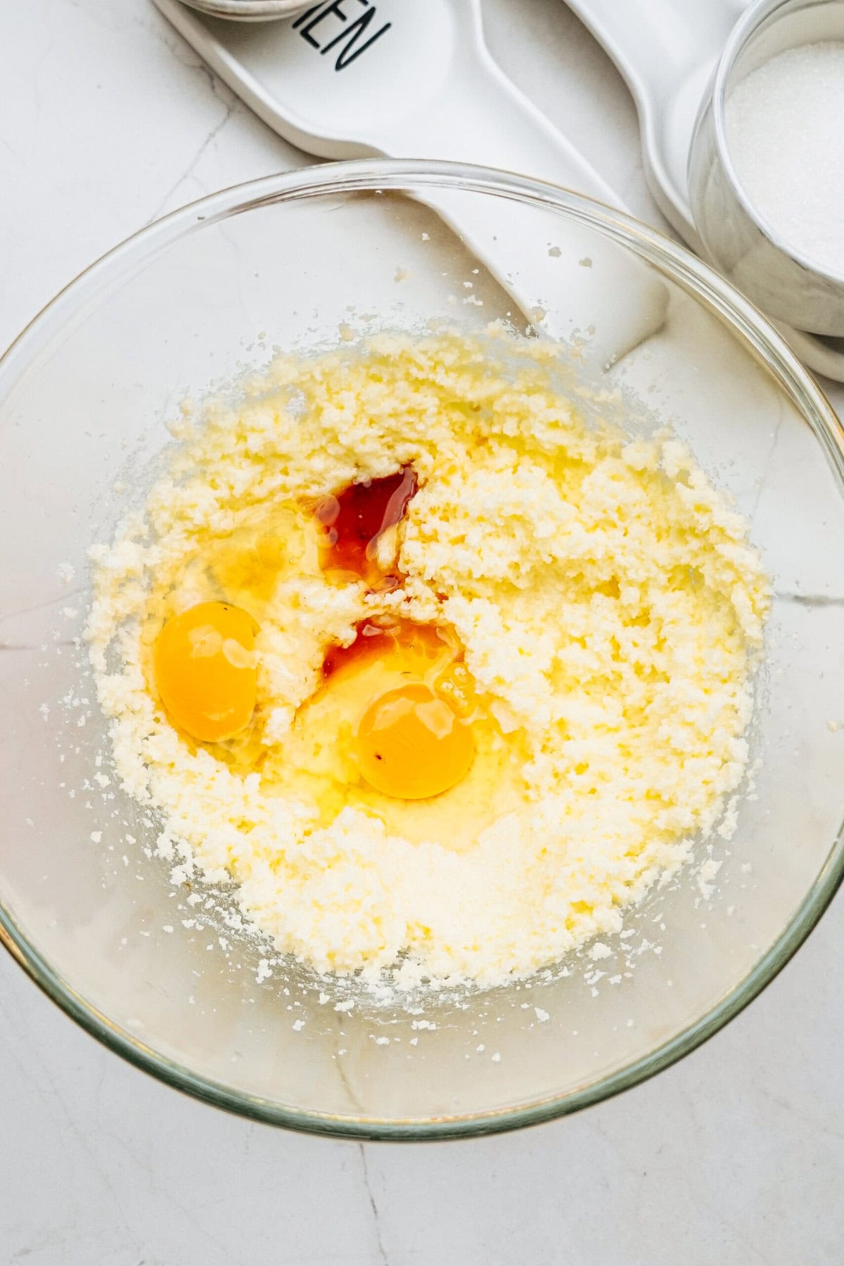 A mixing bowl containing butter, sugar, two raw eggs, and vanilla extract, with measuring spoons and a cup nearby on a white countertop.