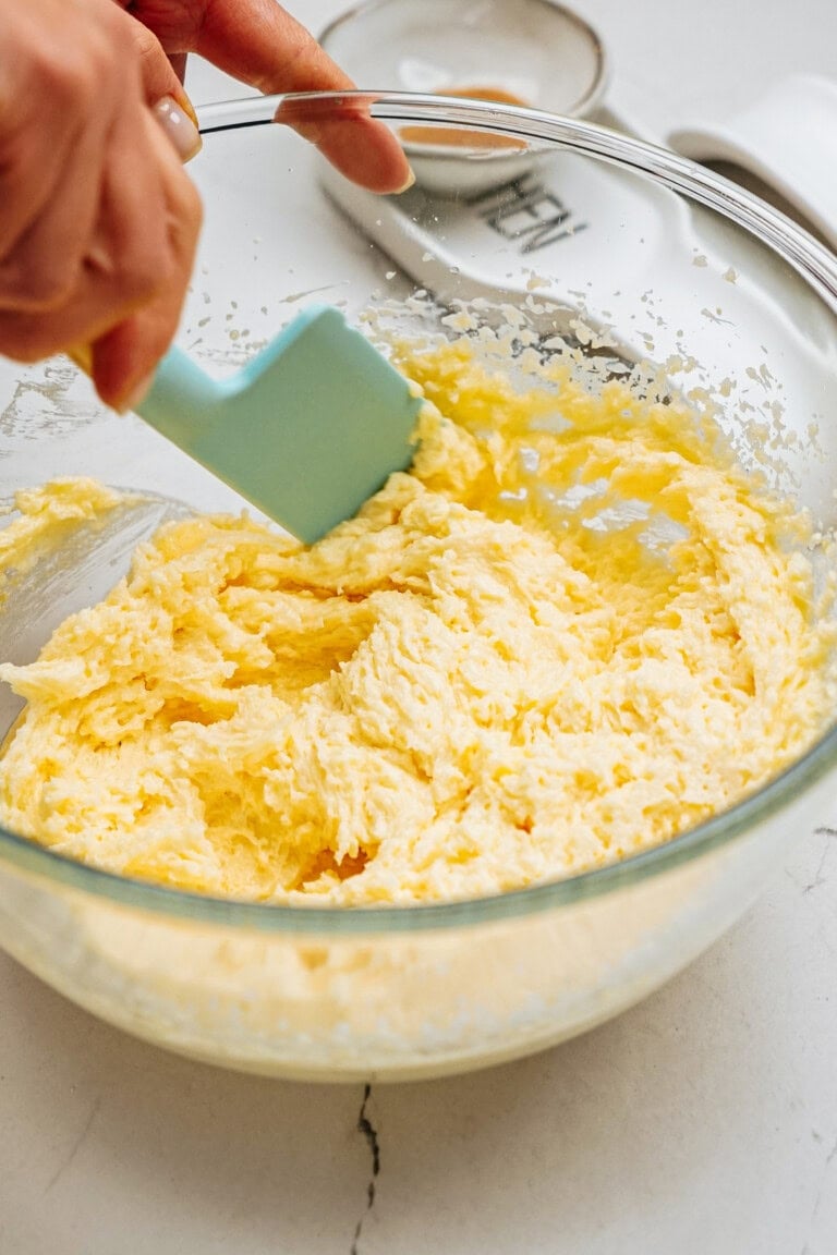 A hand is mixing yellow batter in a clear glass bowl with a blue spatula on a kitchen countertop.