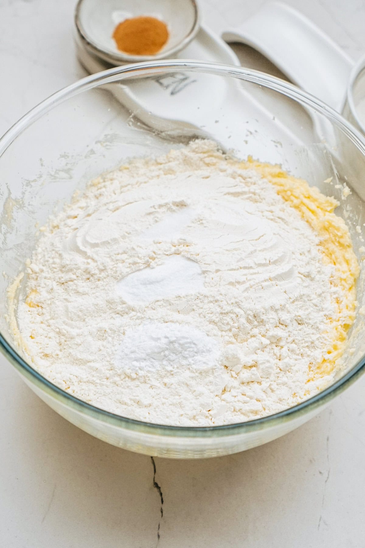 A glass bowl with flour, baking powder, and other dry ingredients. A small bowl with cinnamon and white measuring cups are in the background on a marble surface.