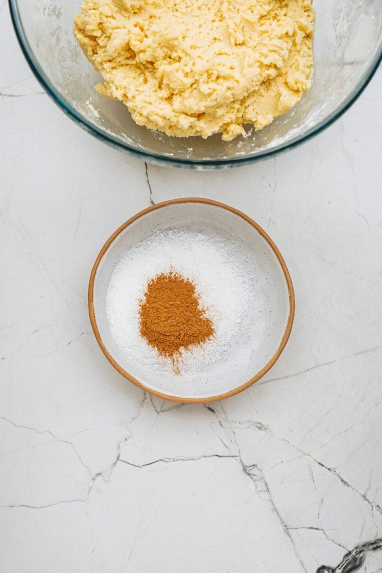 A bowl of sugar with cinnamon on top, next to a glass bowl of dough on a marble countertop.