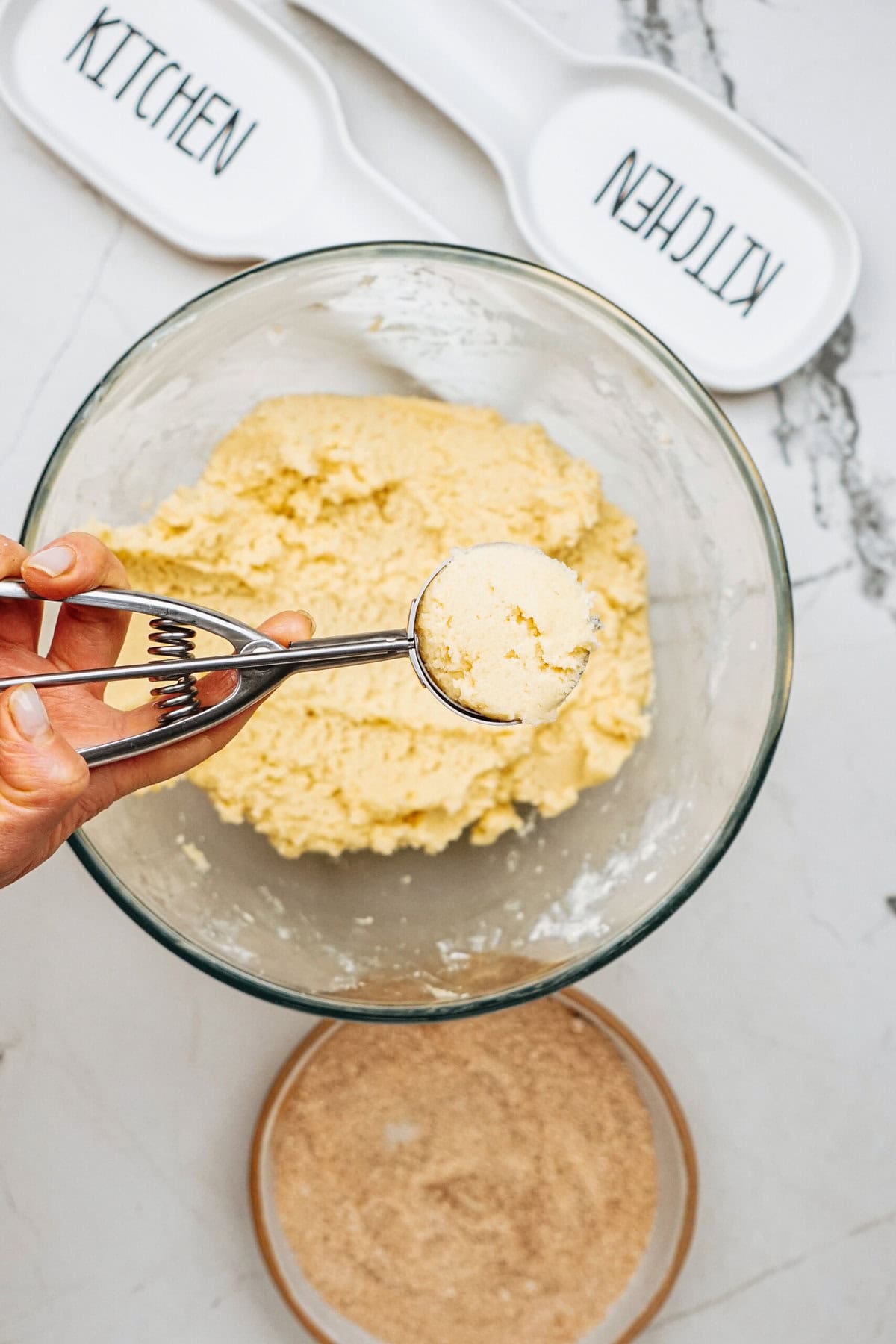 A hand holds a cookie scoop with dough above a bowl of more dough. Two plates labeled "KITCHEN" and a bowl of brown sugar are in the background.