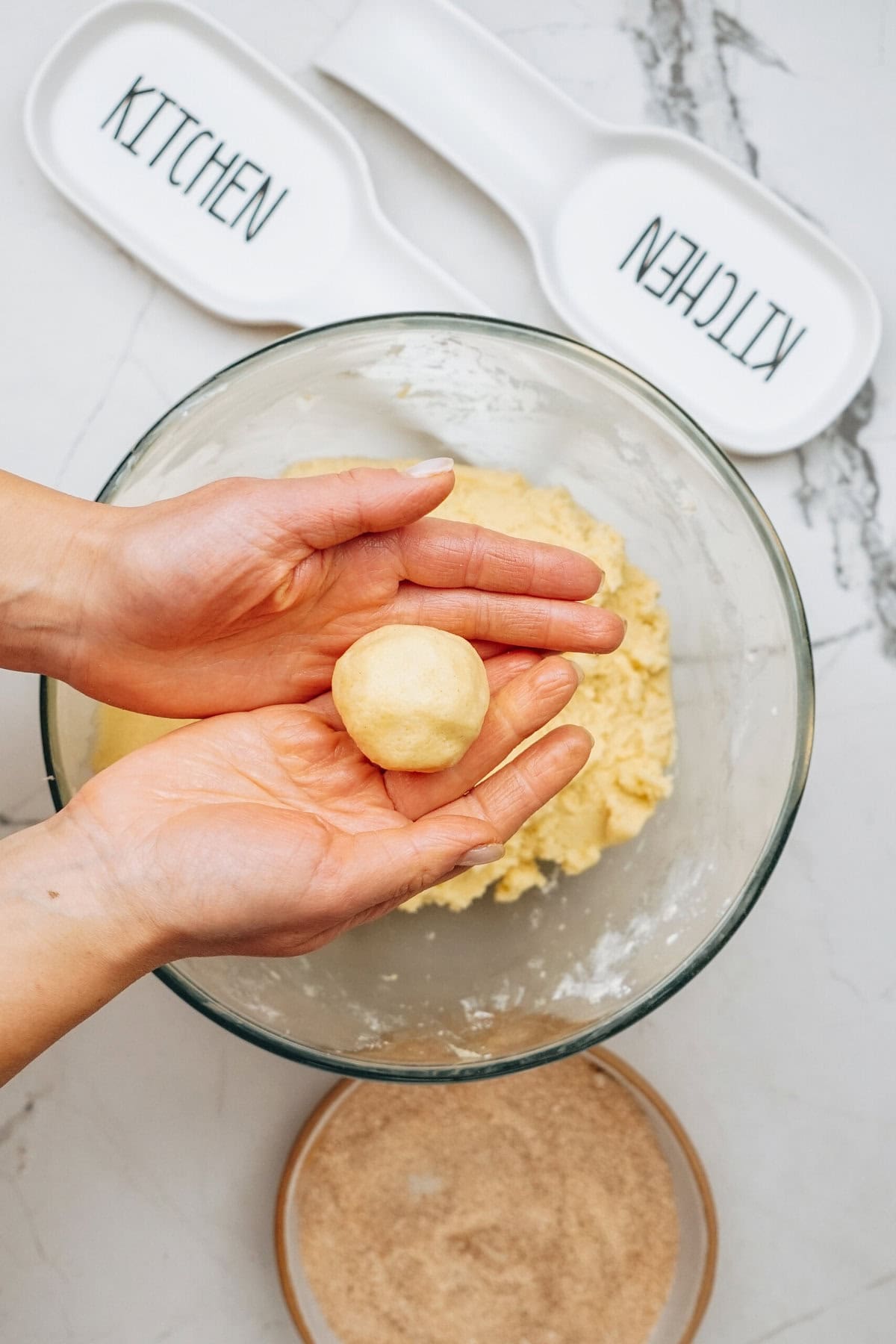 Hands holding a ball of dough over a glass bowl, with baking trays labeled "KITCHEN" in the background.