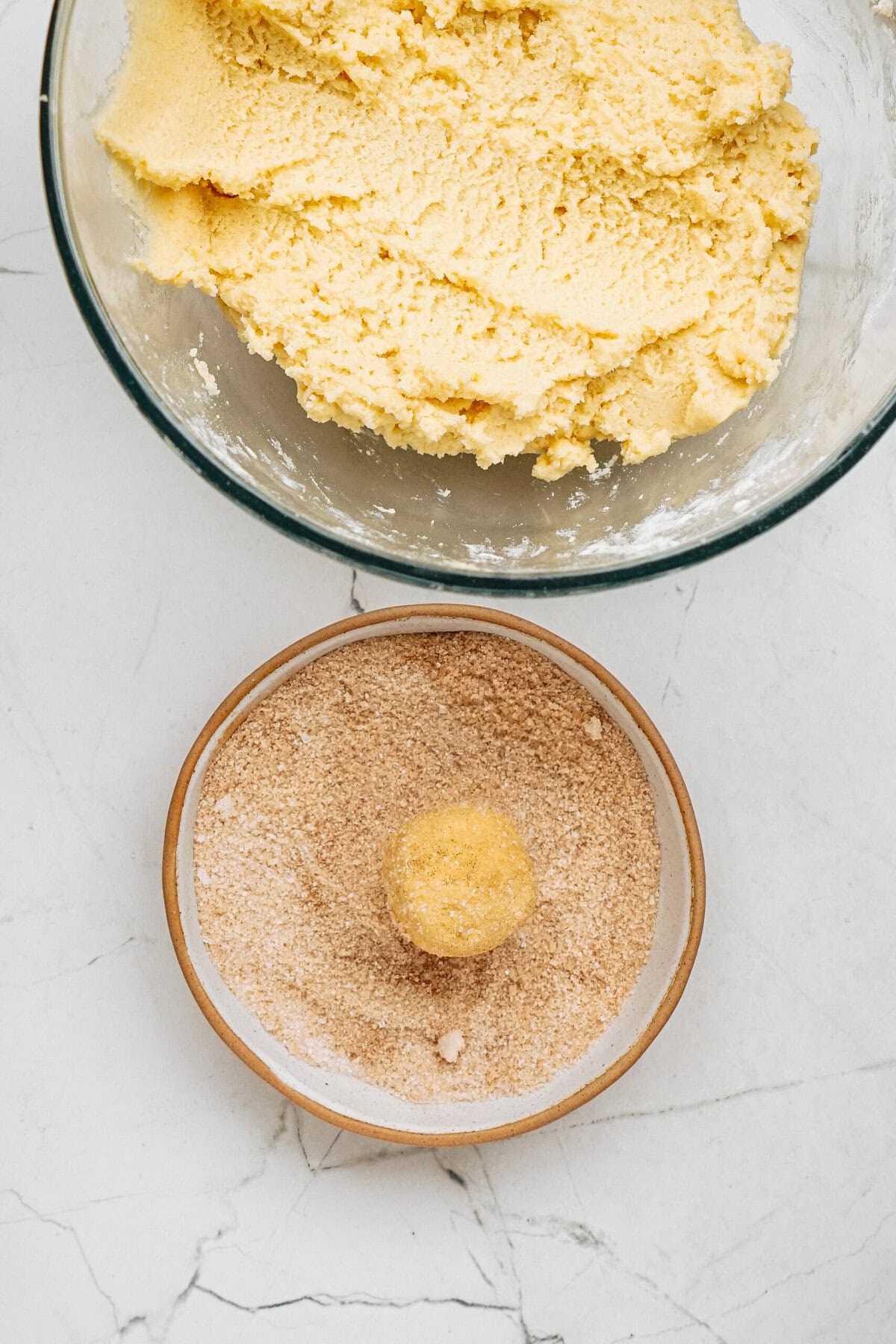 A ball of cookie dough sits on a plate of cinnamon sugar, next to a bowl of unformed dough, on a marble countertop.