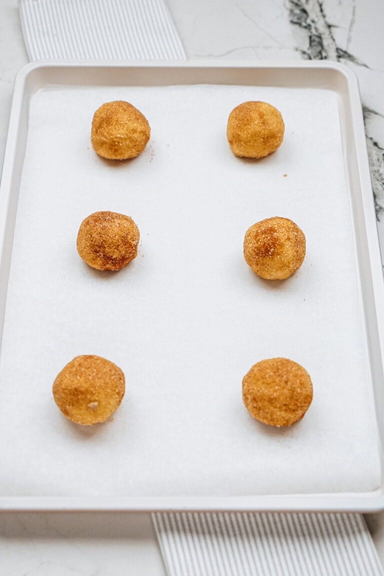 Six round dough balls coated in cinnamon sugar are arranged uniformly on a parchment-lined baking tray.