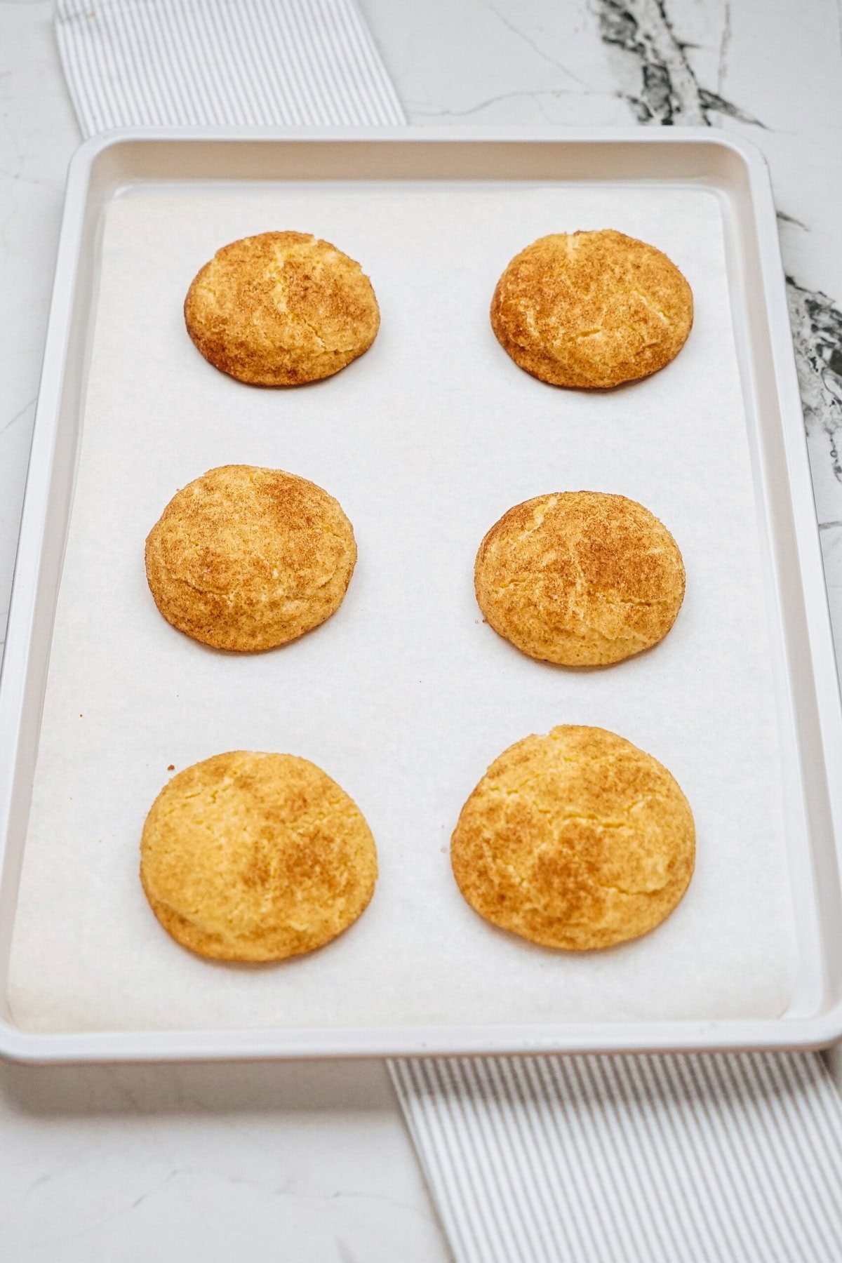 A baking sheet holds six golden-brown cookies on parchment paper, placed on a marble countertop.