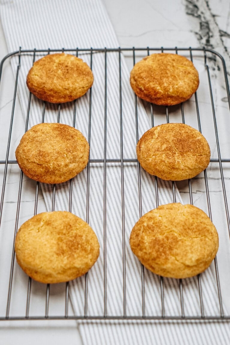 Six golden brown cookies cooling on a wire rack, placed on a white and gray striped cloth on a marble surface.