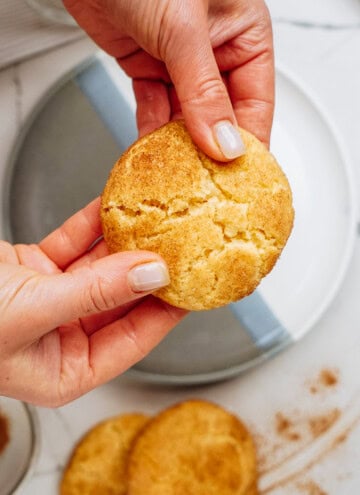 Hands breaking a round, snickerdoodle cookie over a plate, with more cookies and cinnamon nearby.