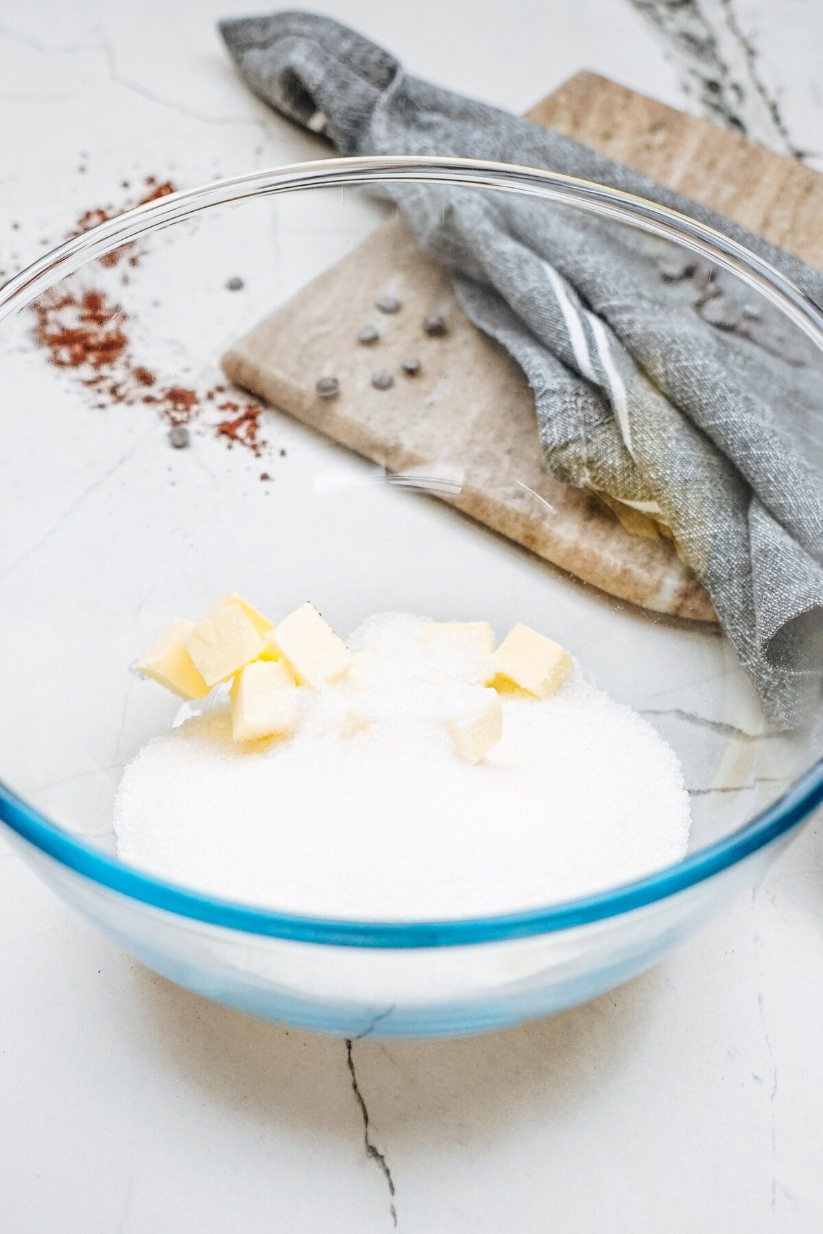 A glass bowl with sugar and butter cubes on a marble countertop. A wooden board with a dark cloth and scattered spices is in the background.