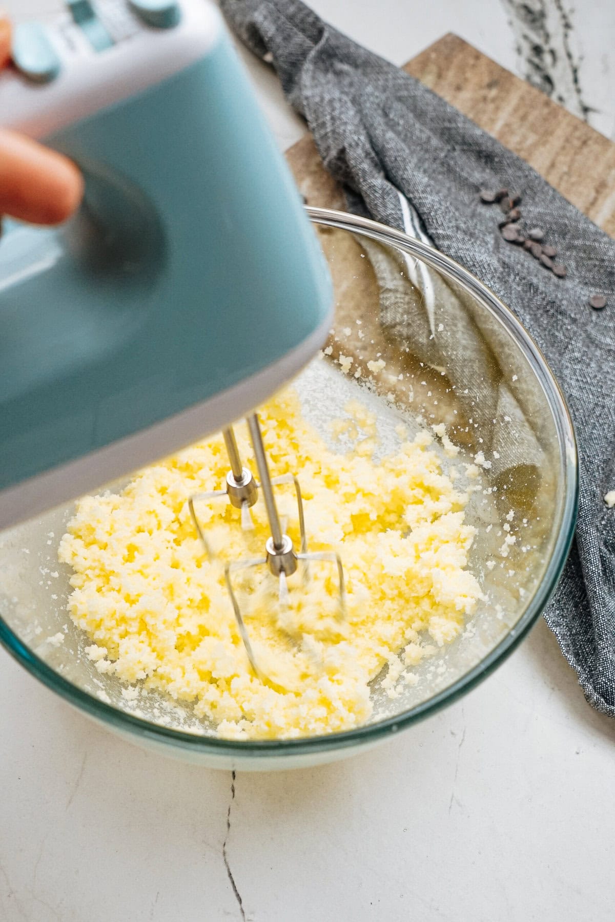 A hand mixer blends butter and sugar in a glass bowl on a marble countertop, with a grey kitchen towel and a cutting board nearby.