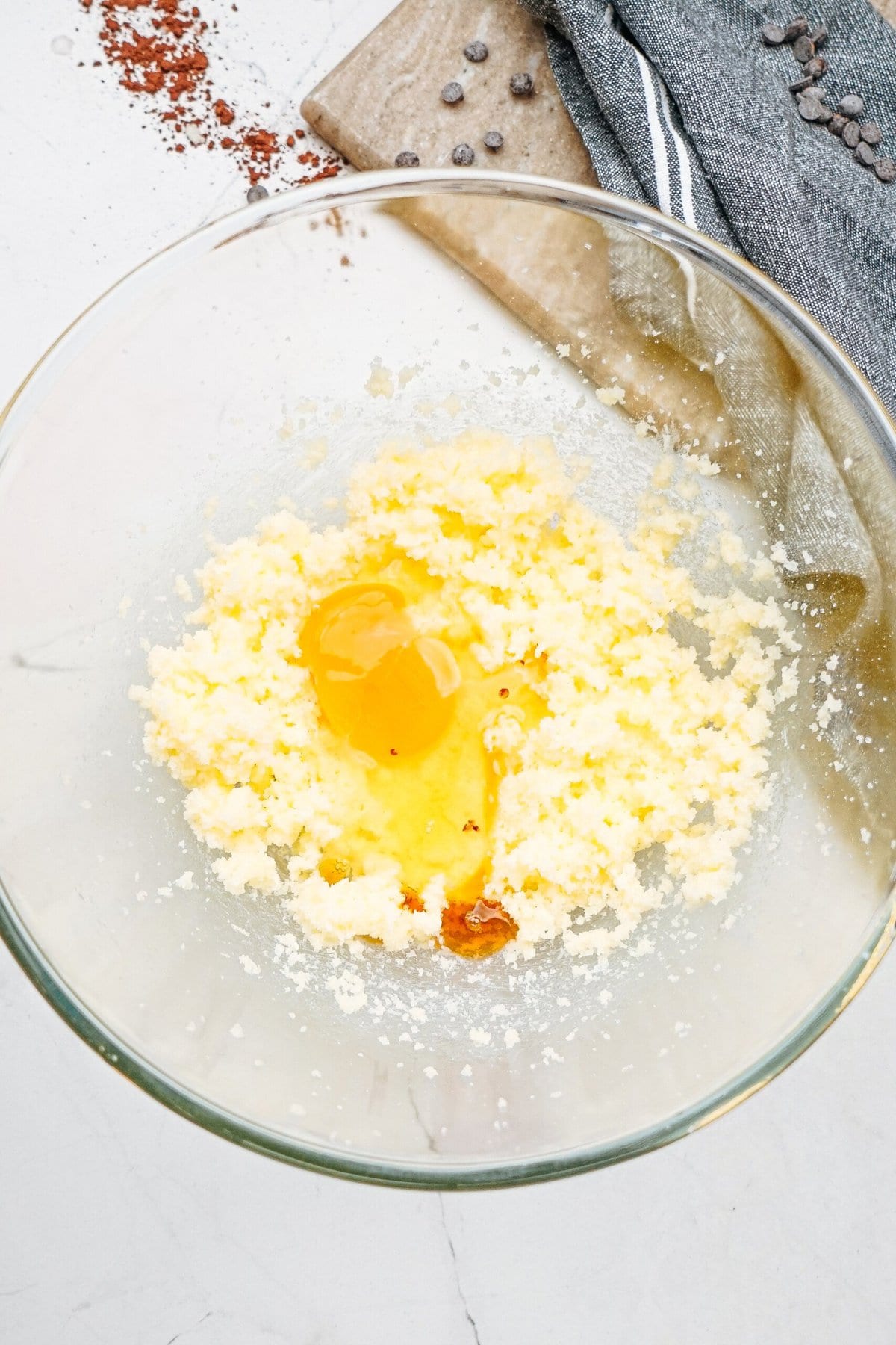 A glass bowl with creamed butter, sugar, and a cracked egg on a white surface, with a wooden board and cloth in the background.