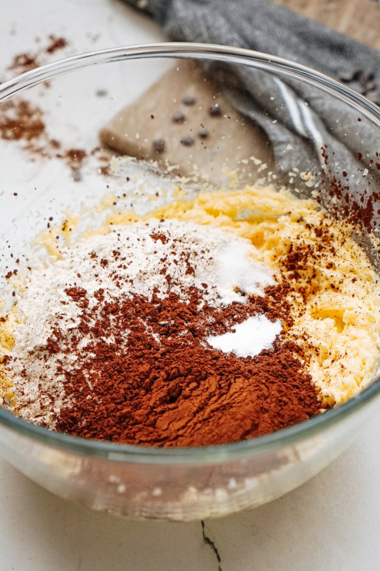 Mixing bowl with flour, cocoa powder, sugar, and butter on a countertop, with a gray cloth in the background.