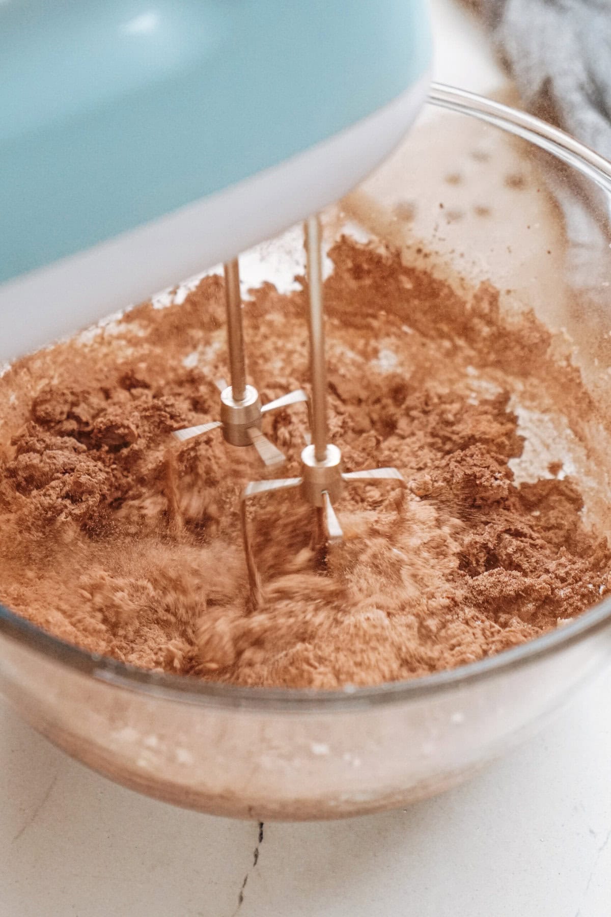 A hand mixer blends cocoa powder and other ingredients in a clear glass bowl.