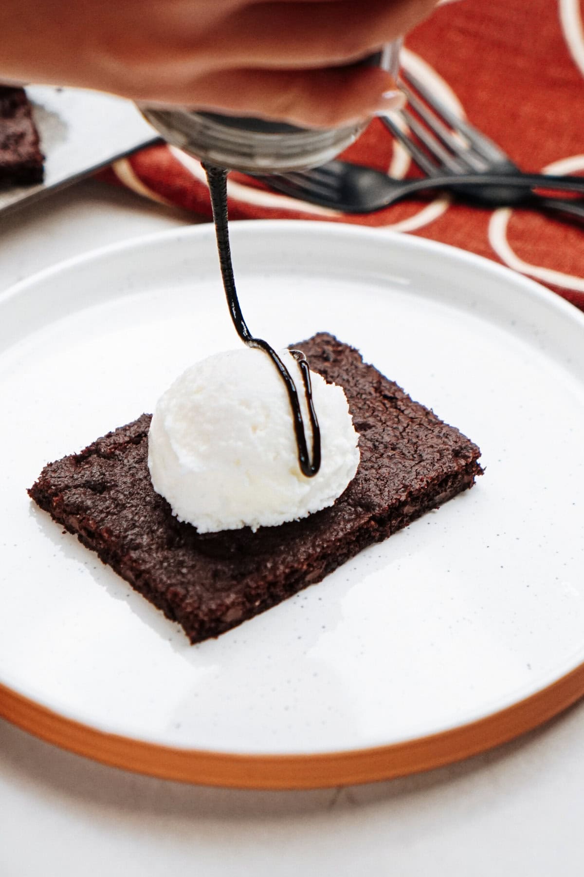 A scoop of ice cream sits on a rectangular brownie, with chocolate syrup being drizzled on top, all on a white plate. A red cloth and forks are in the background.