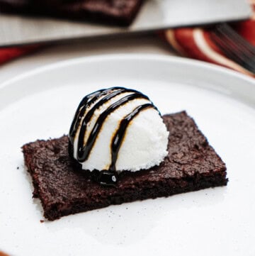 Brownie topped with a scoop of vanilla ice cream and chocolate syrup on a white plate. A tray with more brownies is visible in the background.