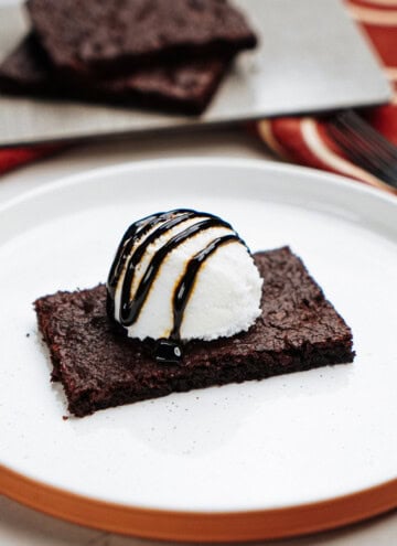 Brownie topped with a scoop of vanilla ice cream and chocolate syrup on a white plate. A tray with more brownies is visible in the background.