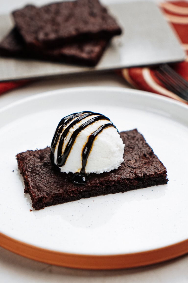 Brownie topped with a scoop of vanilla ice cream and chocolate syrup on a white plate. A tray with more brownies is visible in the background.