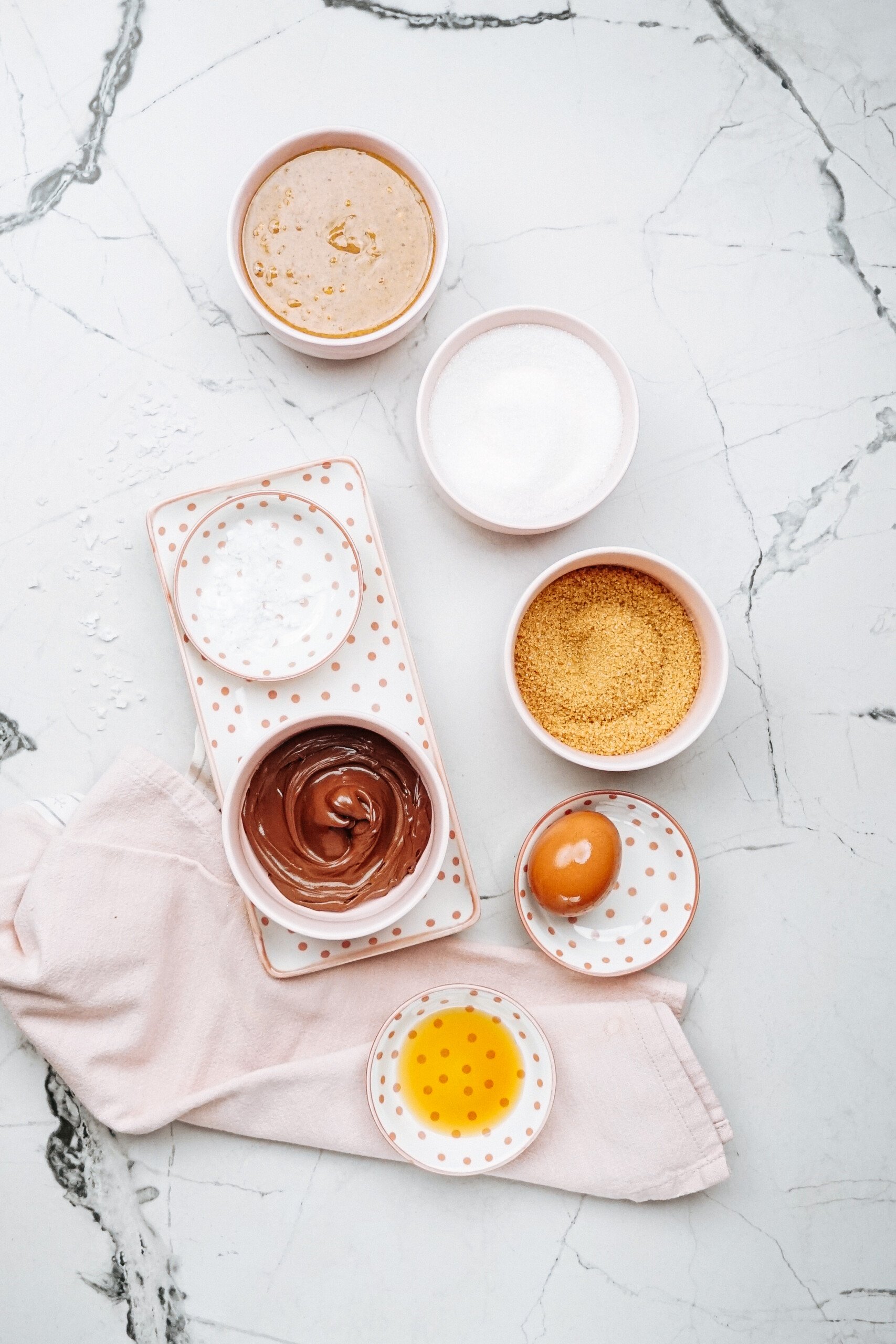 Ingredients for Peanut Butter Thumbprint Cookies are laid out on a marble countertop, featuring bowls of peanut butter, sugar, brown sugar, salt, and chocolate spread. An egg and a small dish of vanilla extract complete the delightful ensemble for these irresistible treats.