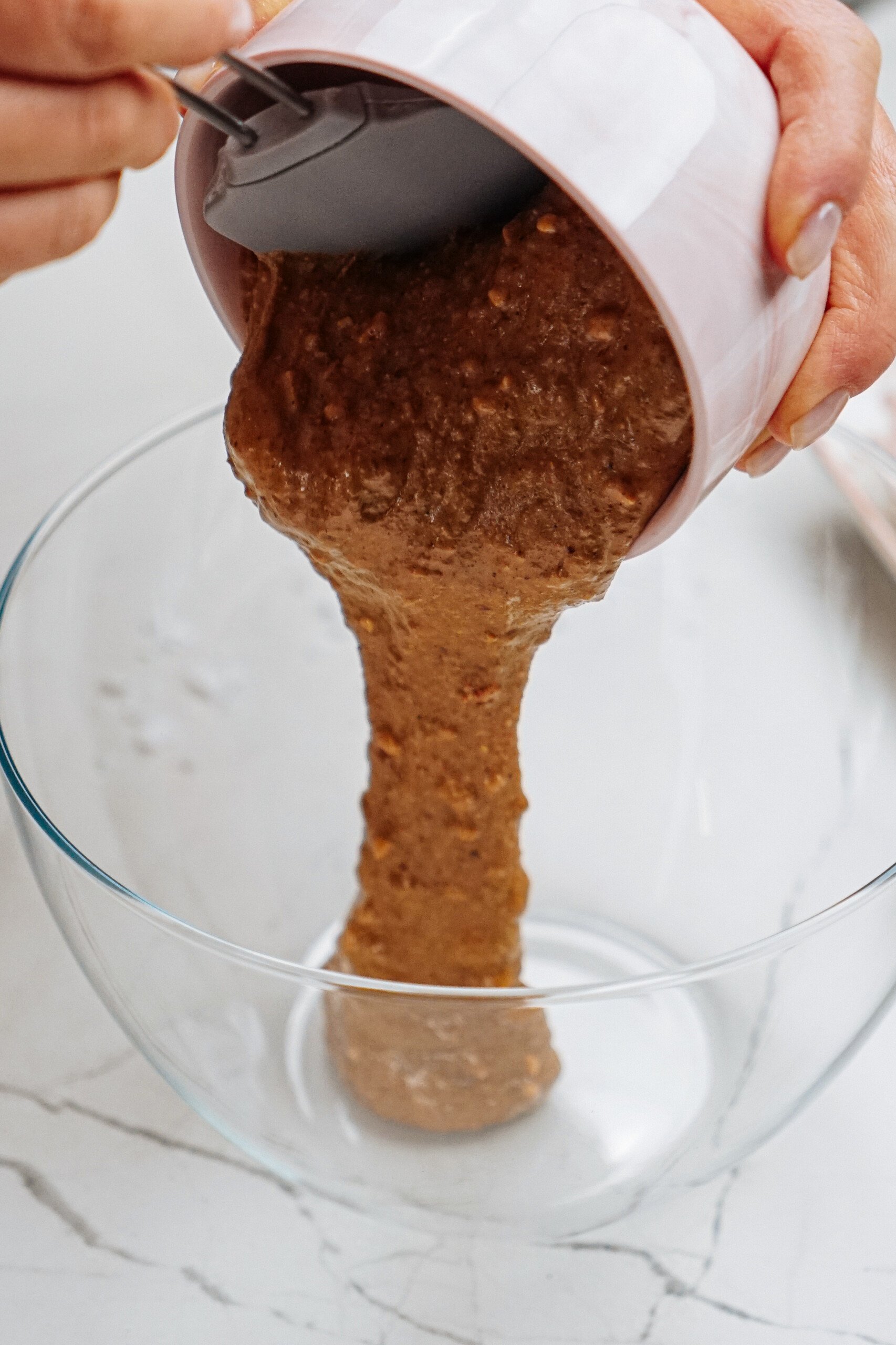 Peanut butter thumbprint cookies start to take shape as batter cascades gracefully from a pink bowl into a clear mixing dish, ready to be transformed into delicious bites of joy.