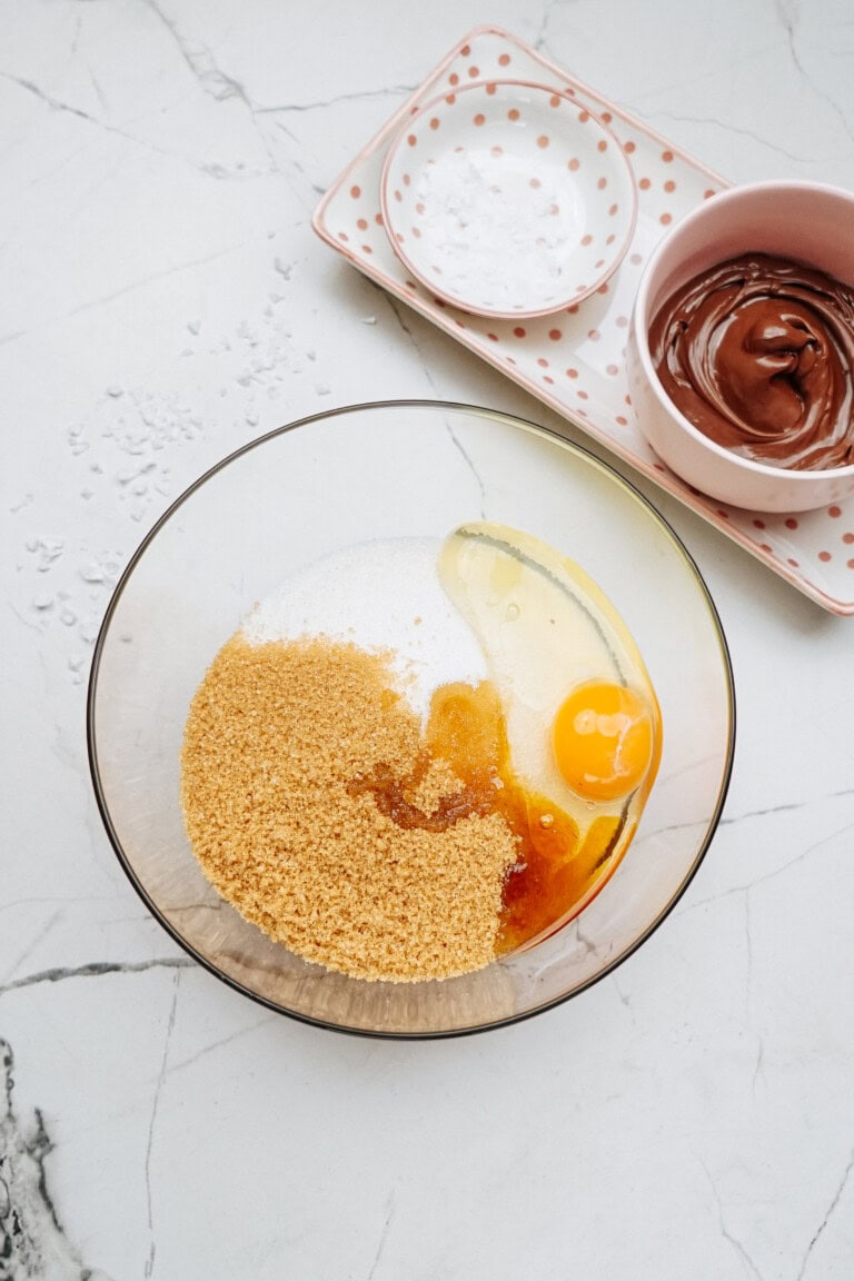 A bowl holds brown sugar, granulated sugar, and an egg, ready to blend into the perfect mix. Nearby, a dish with cocoa powder and a bowl of melted chocolate await, hinting at the rich flavors of Peanut Butter Thumbprint Cookies.