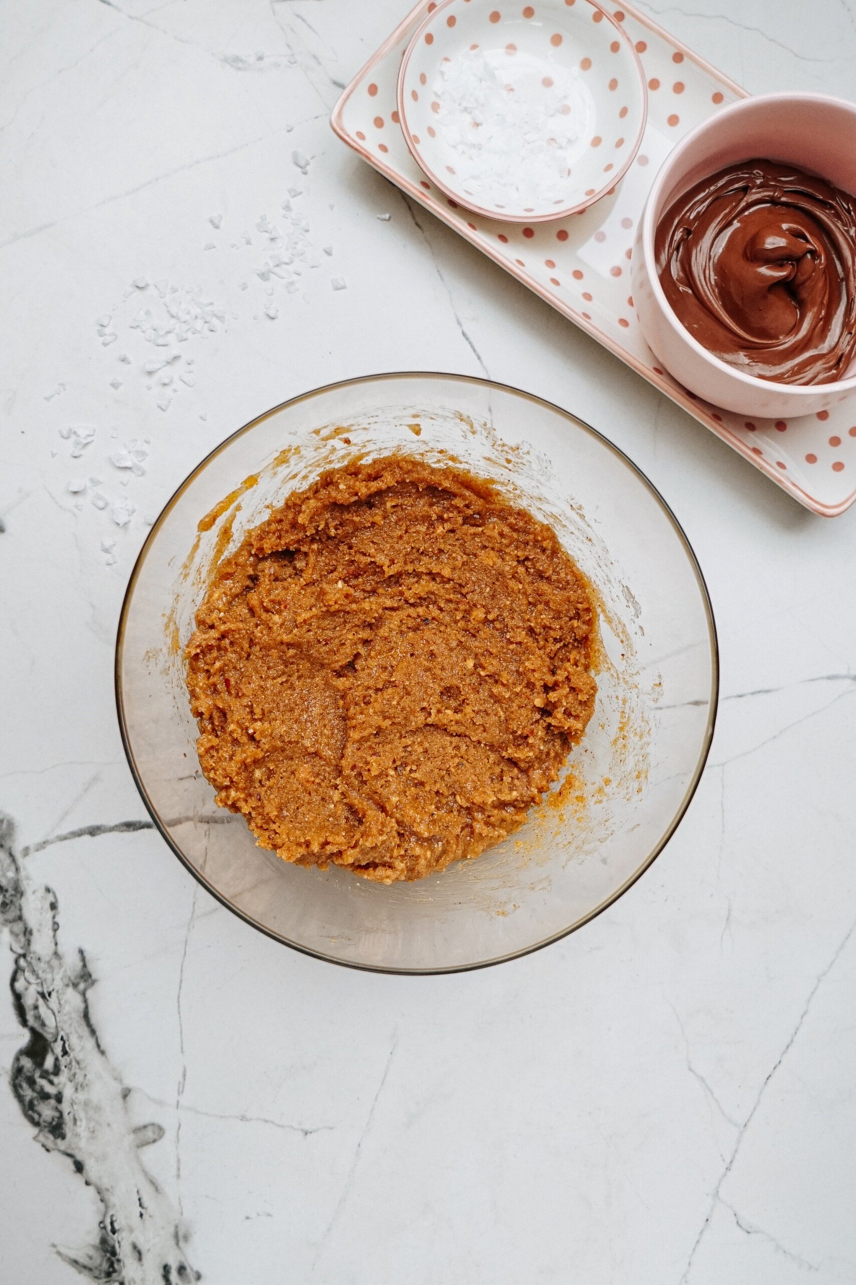 A bowl of cookie dough sits on a marble surface next to a small bowl of melted chocolate, ready to drizzle over peanut butter thumbprint cookies resting on an empty dotted plate.