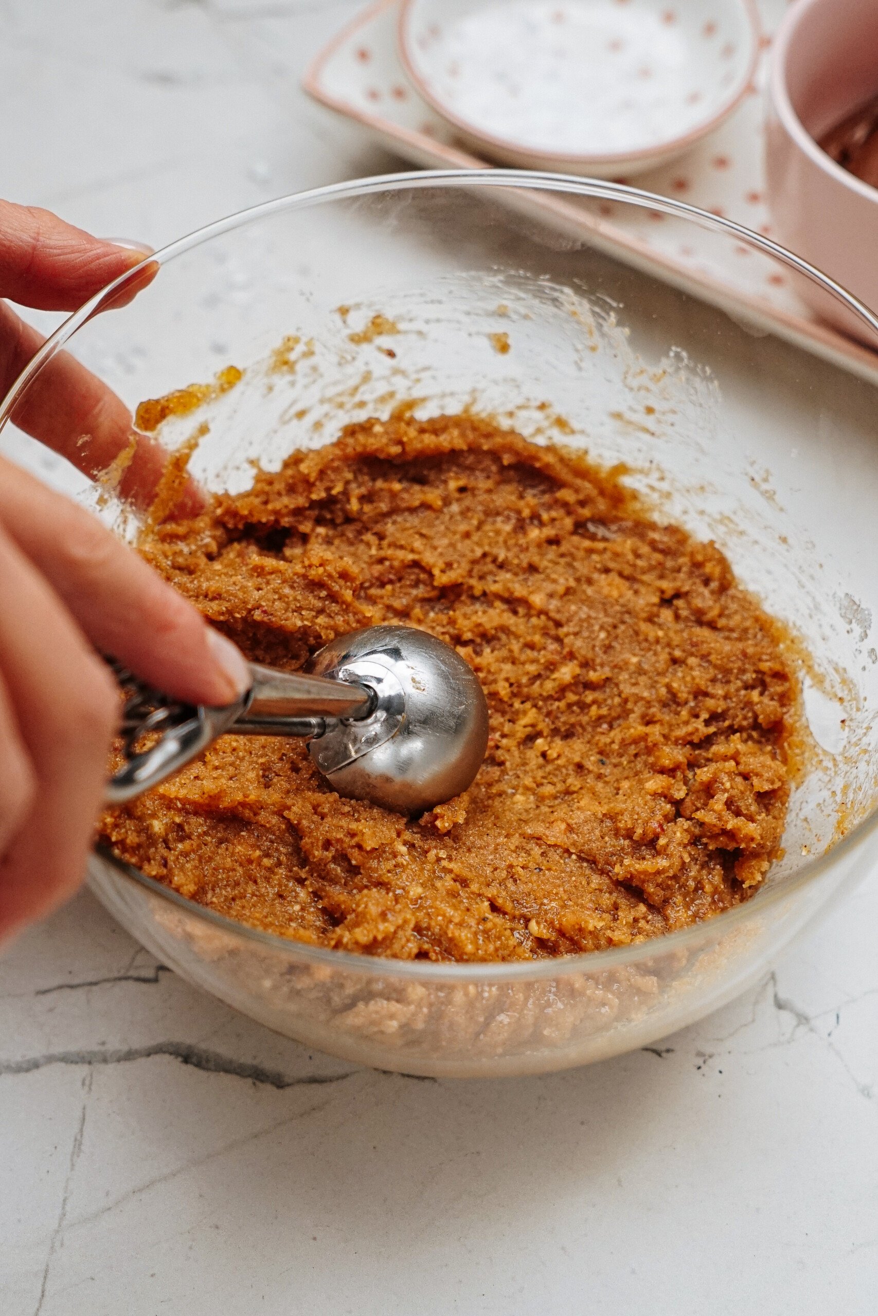 A person uses a scoop to prepare cookie dough in a glass bowl on a marble countertop, crafting the perfect base for delicious peanut butter thumbprint cookies.