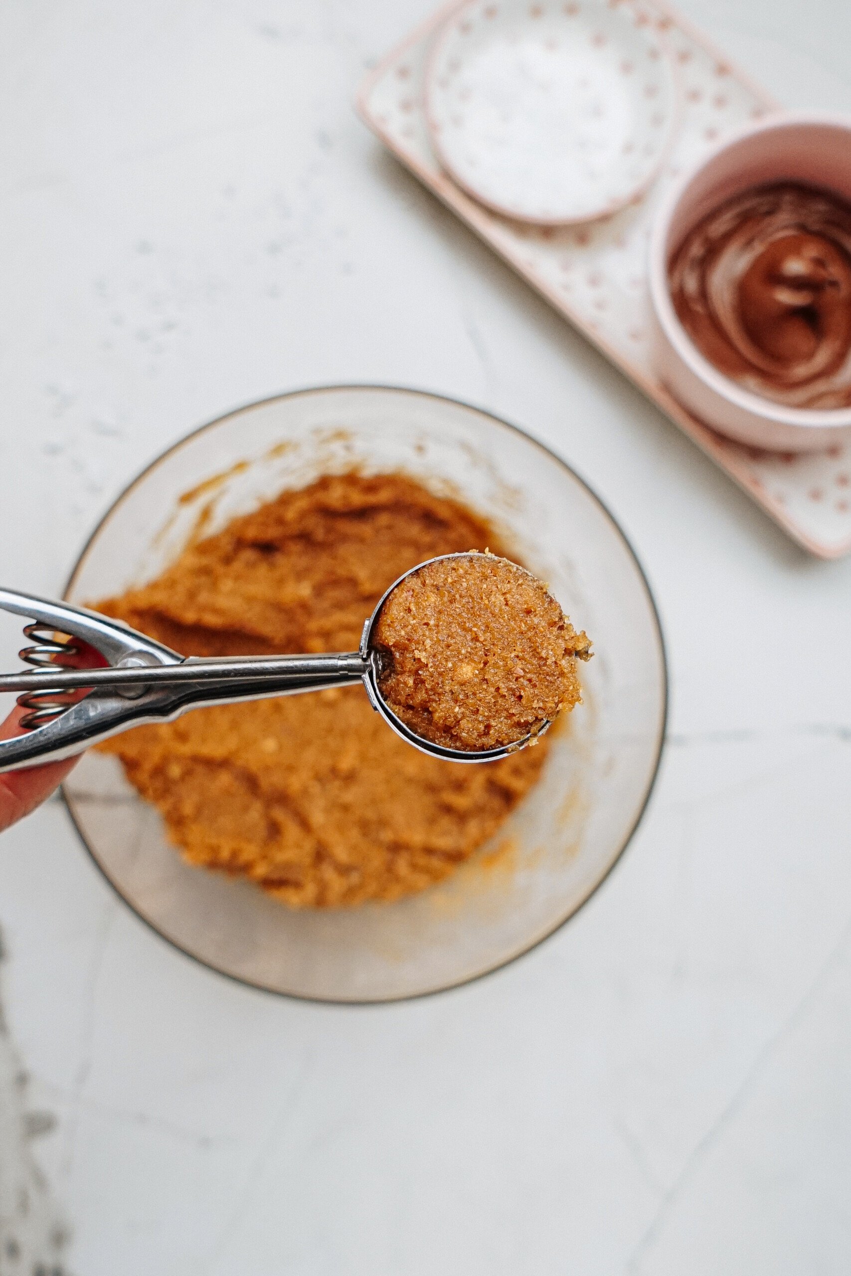 A hand delicately holds a cookie scoop with dough over a bowl, preparing to make delightful Peanut Butter Thumbprint Cookies. In the background, a small dish of chocolate and another dish of white sugar await their turn.