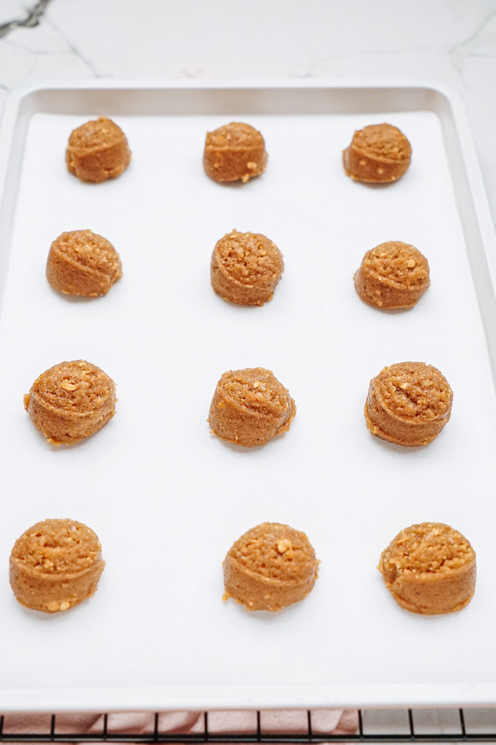 Twelve scoops of cookie dough, ready to become peanut butter thumbprint cookies, are evenly spaced on a parchment-lined baking sheet.