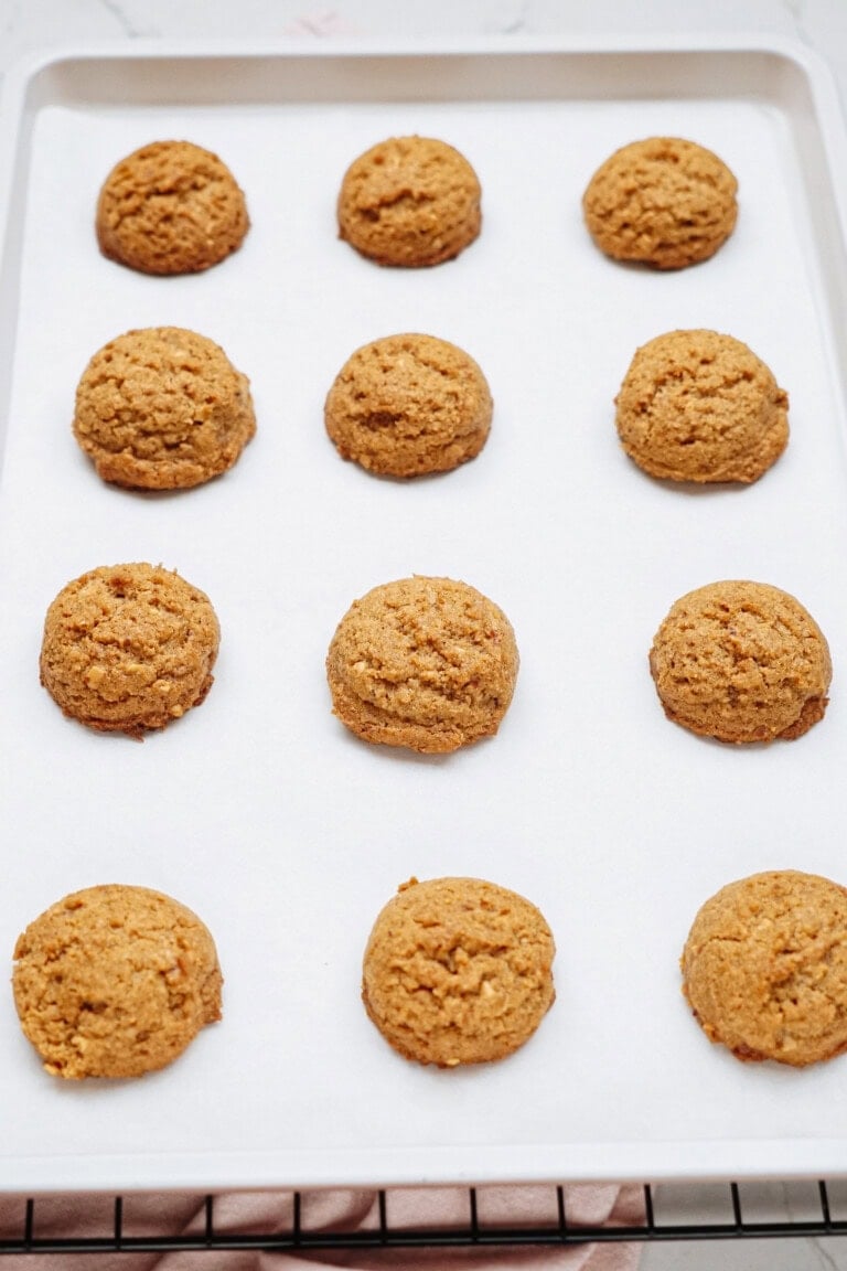 A baking sheet with twelve evenly spaced Peanut Butter Thumbprint Cookies on parchment paper, cooling on a wire rack.