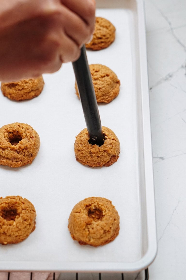 A hand skillfully uses a tool to form wells in peanut butter thumbprint cookies on a baking sheet lined with parchment paper.