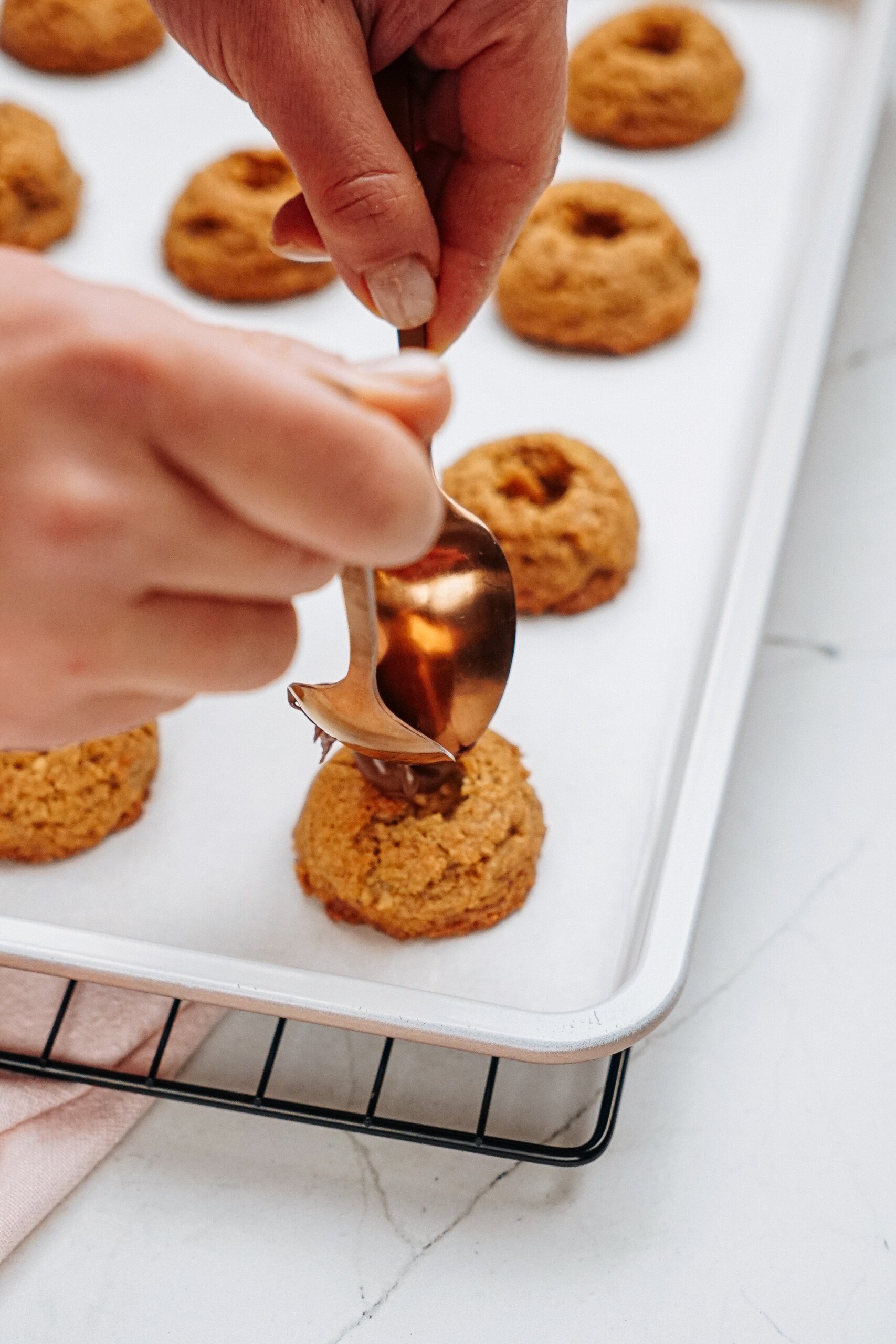 A person using a spoon to fill Peanut Butter Thumbprint Cookies on a baking sheet with chocolate.