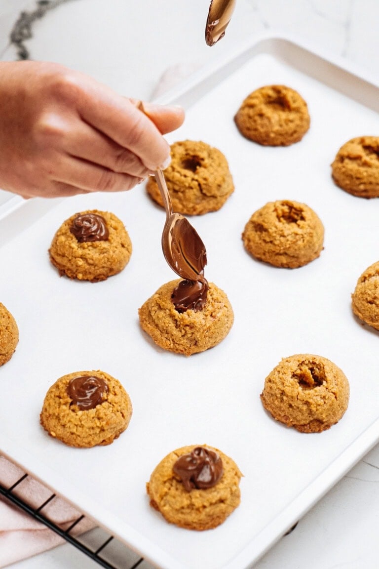 A hand carefully adds chocolate to Peanut Butter Thumbprint Cookies on a baking tray lined with parchment paper.