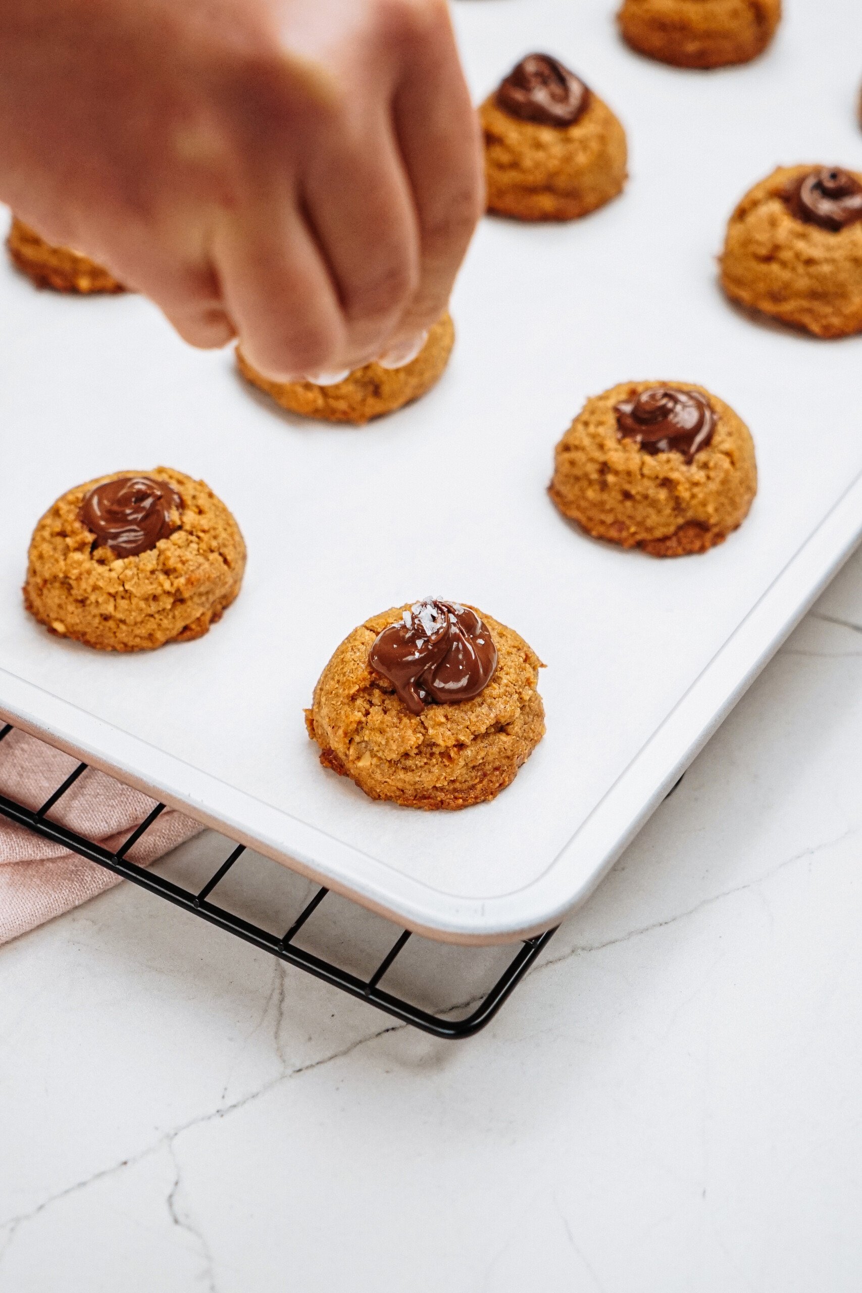 A hand places peanut butter thumbprint cookie dough on a baking sheet lined with parchment, topped with chocolate, ready for baking.