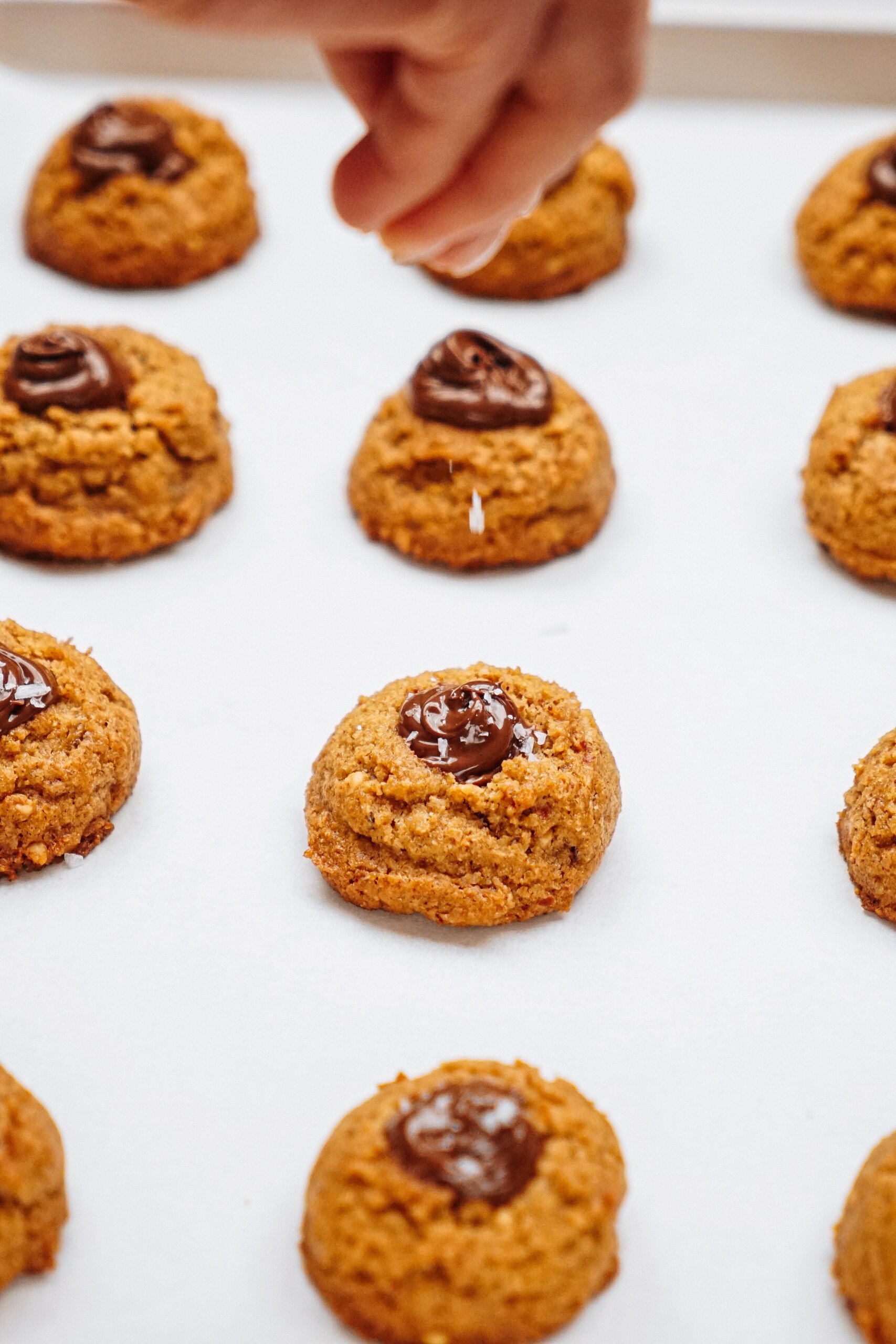 Hand sprinkling salt on freshly baked peanut butter thumbprint cookies topped with chocolate, arranged on parchment paper.