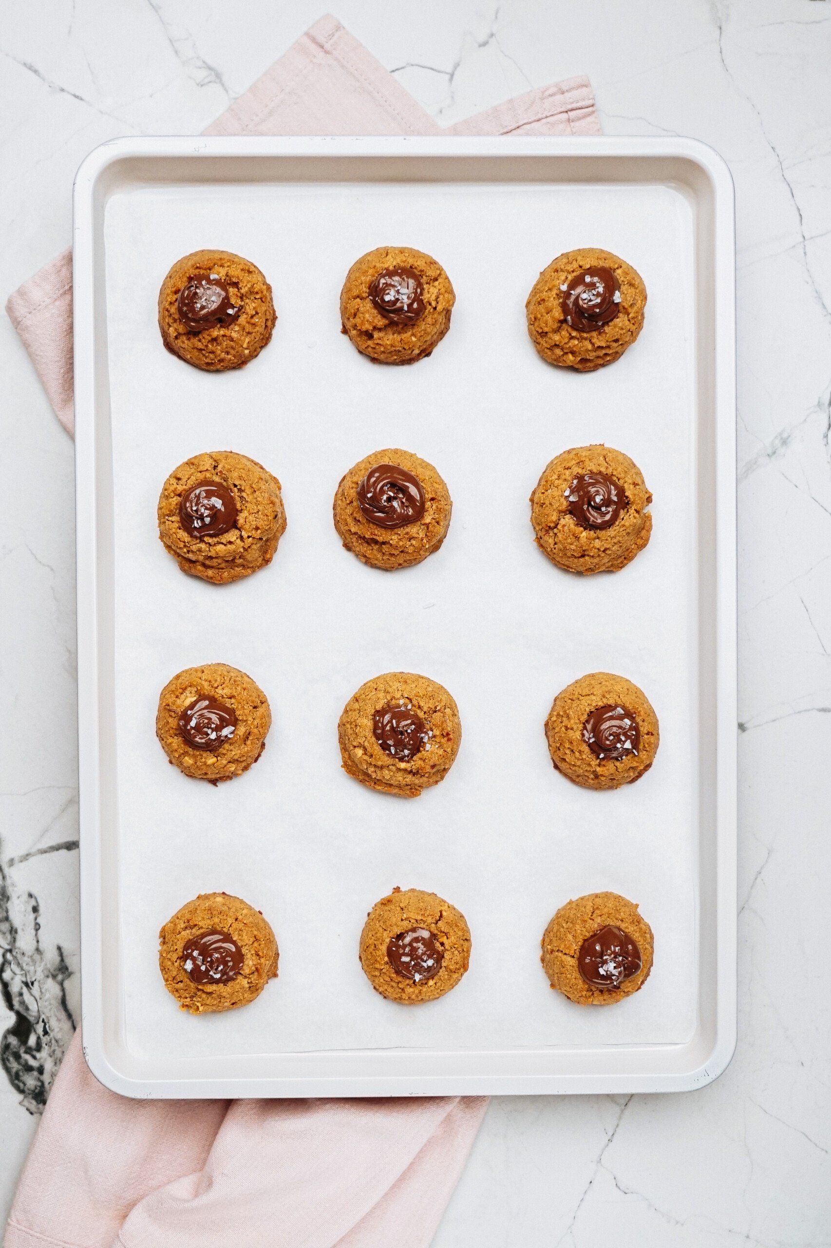 A baking tray with twelve round cookies, each a delightful peanut butter thumbprint cookie topped with a dollop of chocolate, rests on a parchment-lined sheet against a light marble surface.