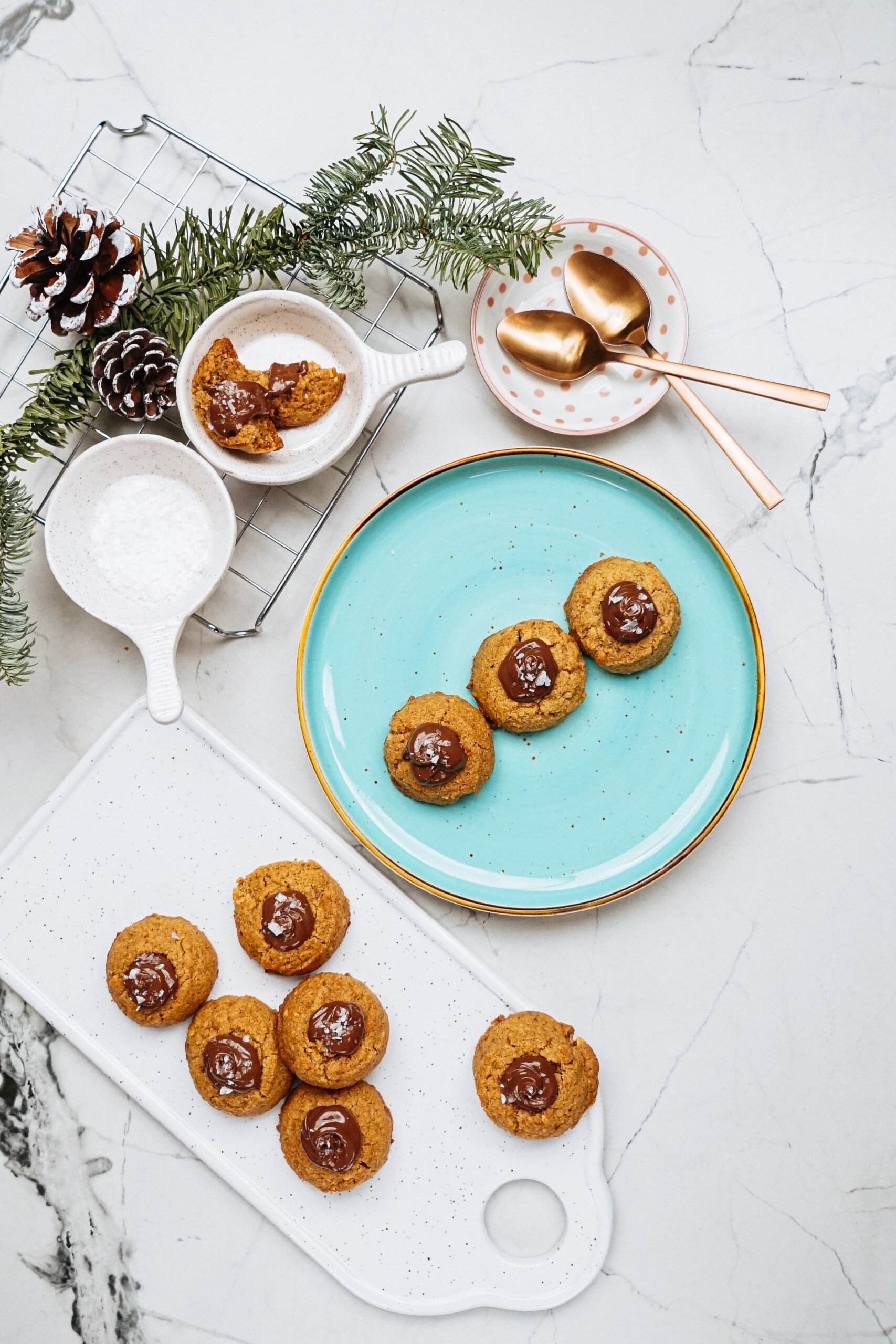 Peanut Butter Thumbprint Cookies with chocolate centers sit on two plates, accompanied by additional cookies, pinecones, and a bowl of sugar on a marble surface. Gold spoons rest elegantly on a polka dot dish.