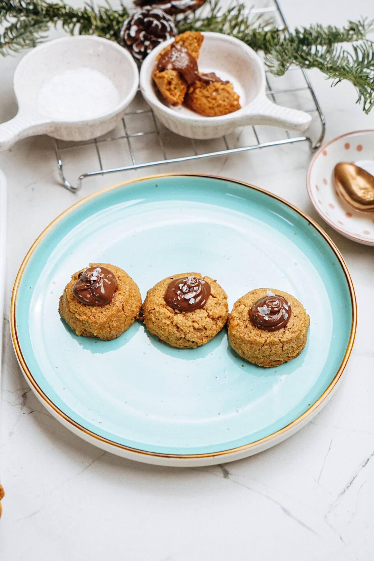 Three peanut butter thumbprint cookies with chocolate centers sit on a teal plate, surrounded by additional cookies and festive decorations.