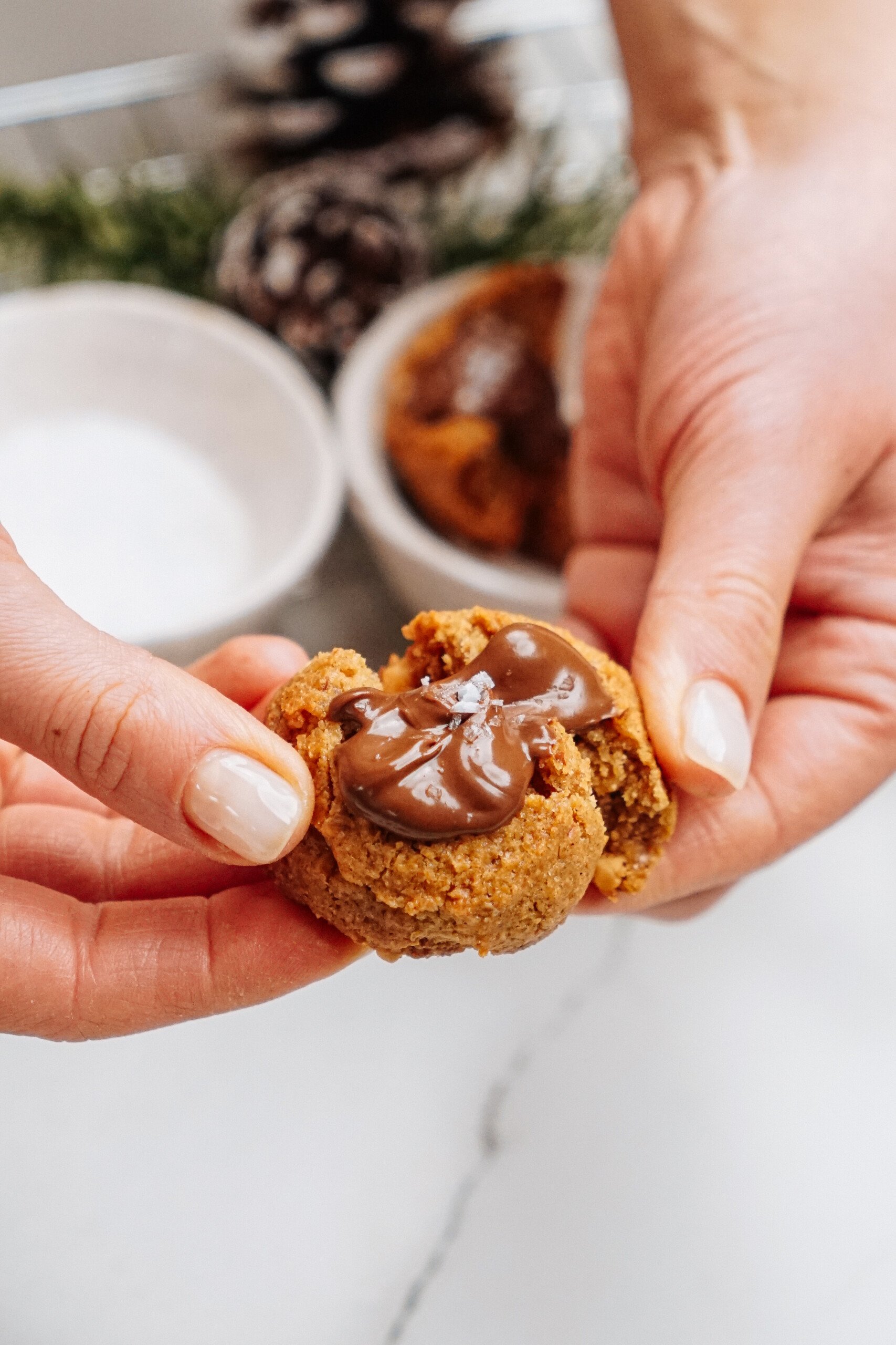 Hands holding a peanut butter thumbprint cookie filled with melted chocolate, with bowls and a pine cone in the background.