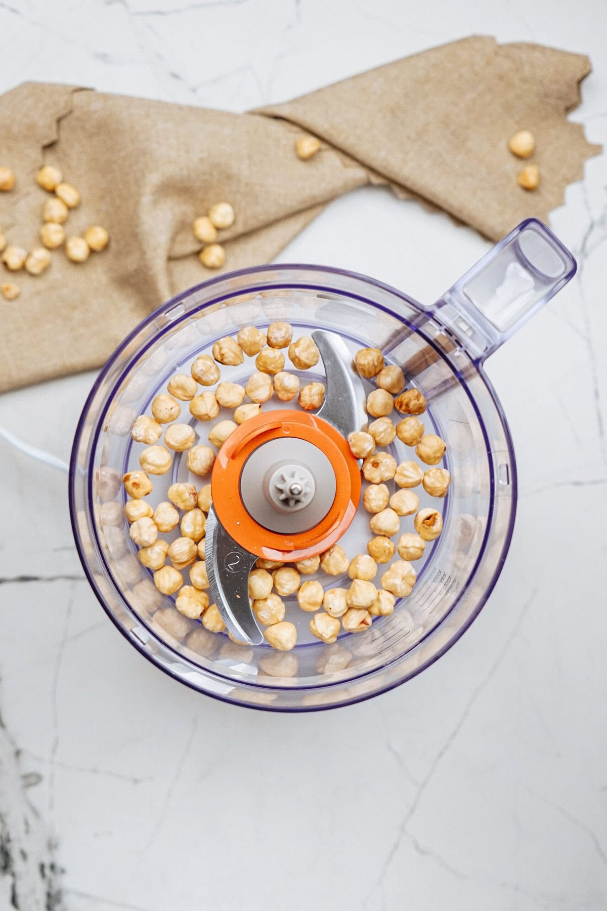 Top view of a food processor with hazelnuts inside on a marble surface, with some hazelnuts scattered and brown fabric in the background.