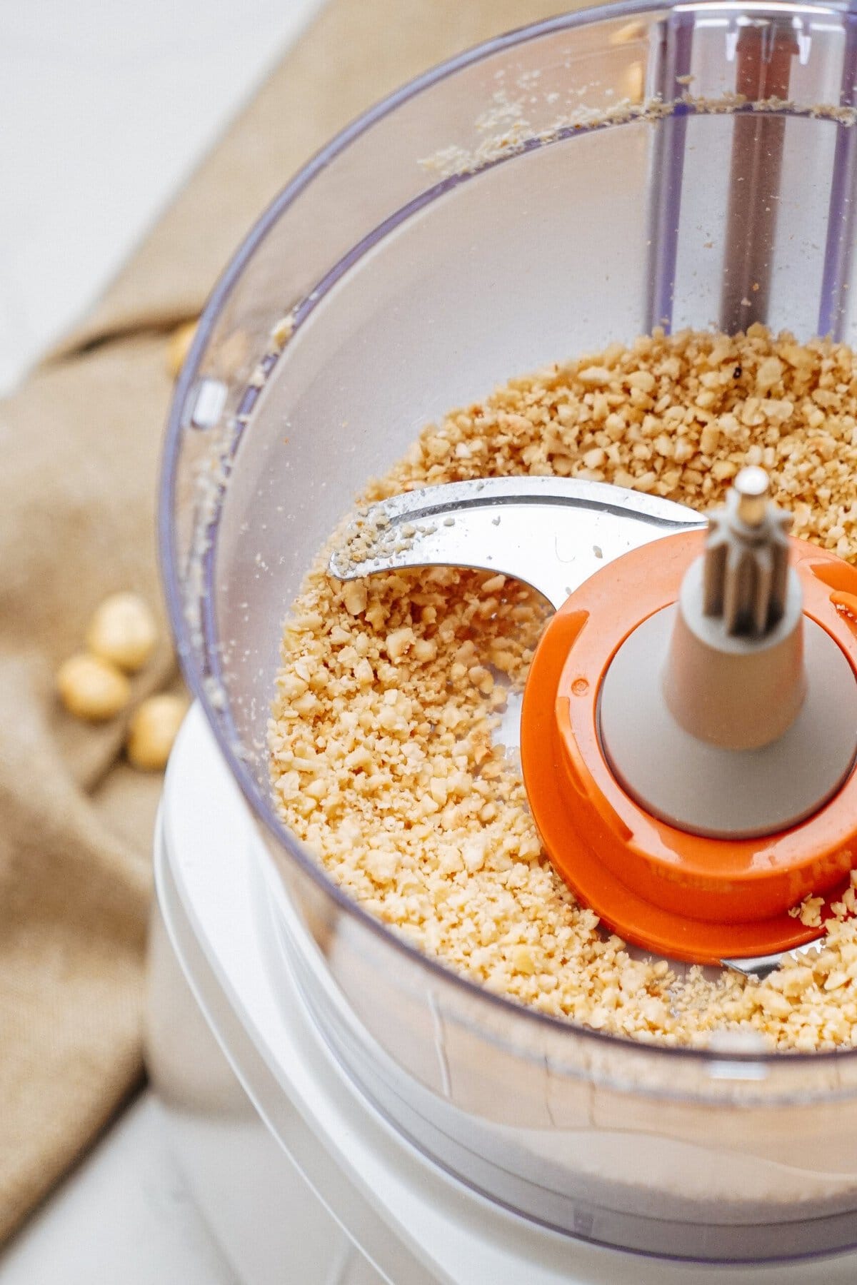 Chopped nuts in a food processor with an orange blade attachment, on a counter with a beige cloth and whole nuts nearby.