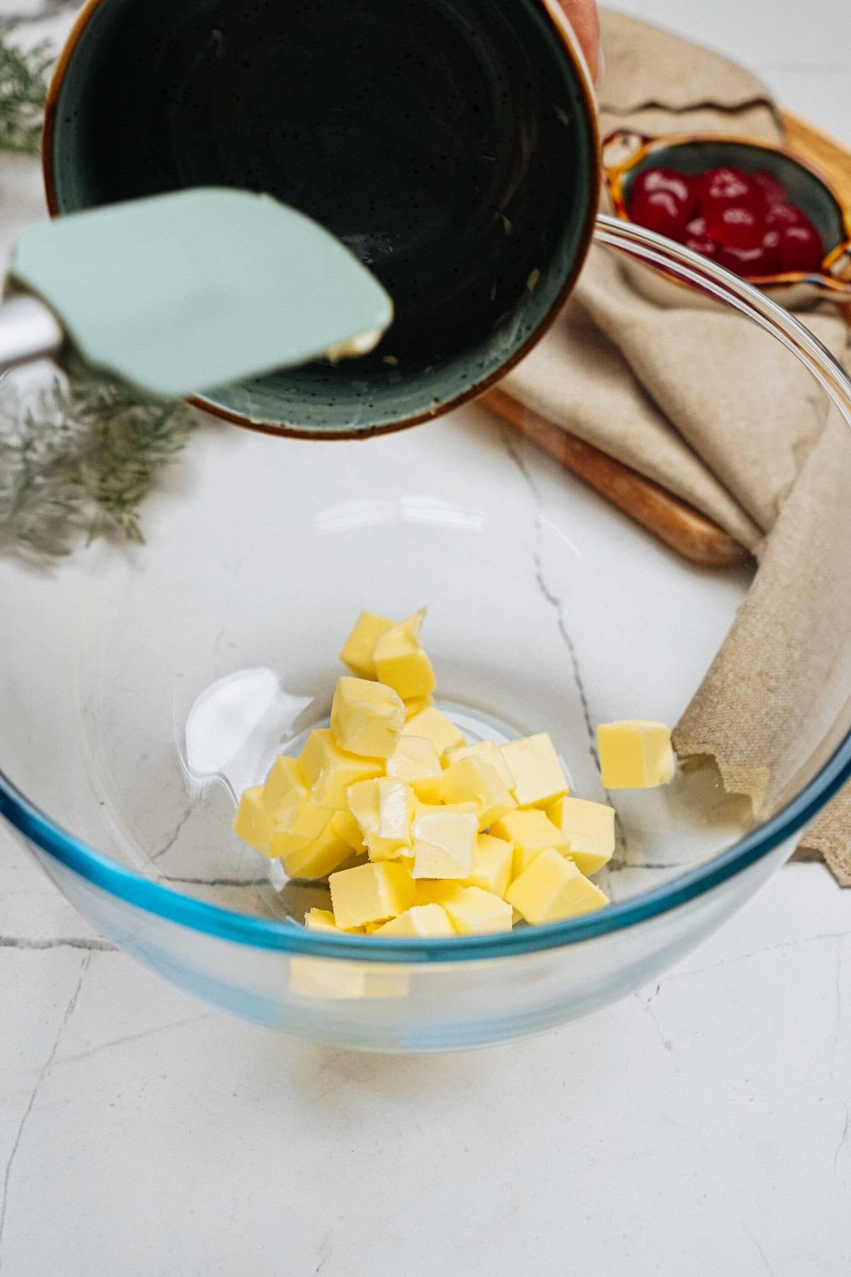 A glass bowl with cubed butter. A spatula and a bowl are visible above. A beige napkin and red berries in a bowl are in the background.