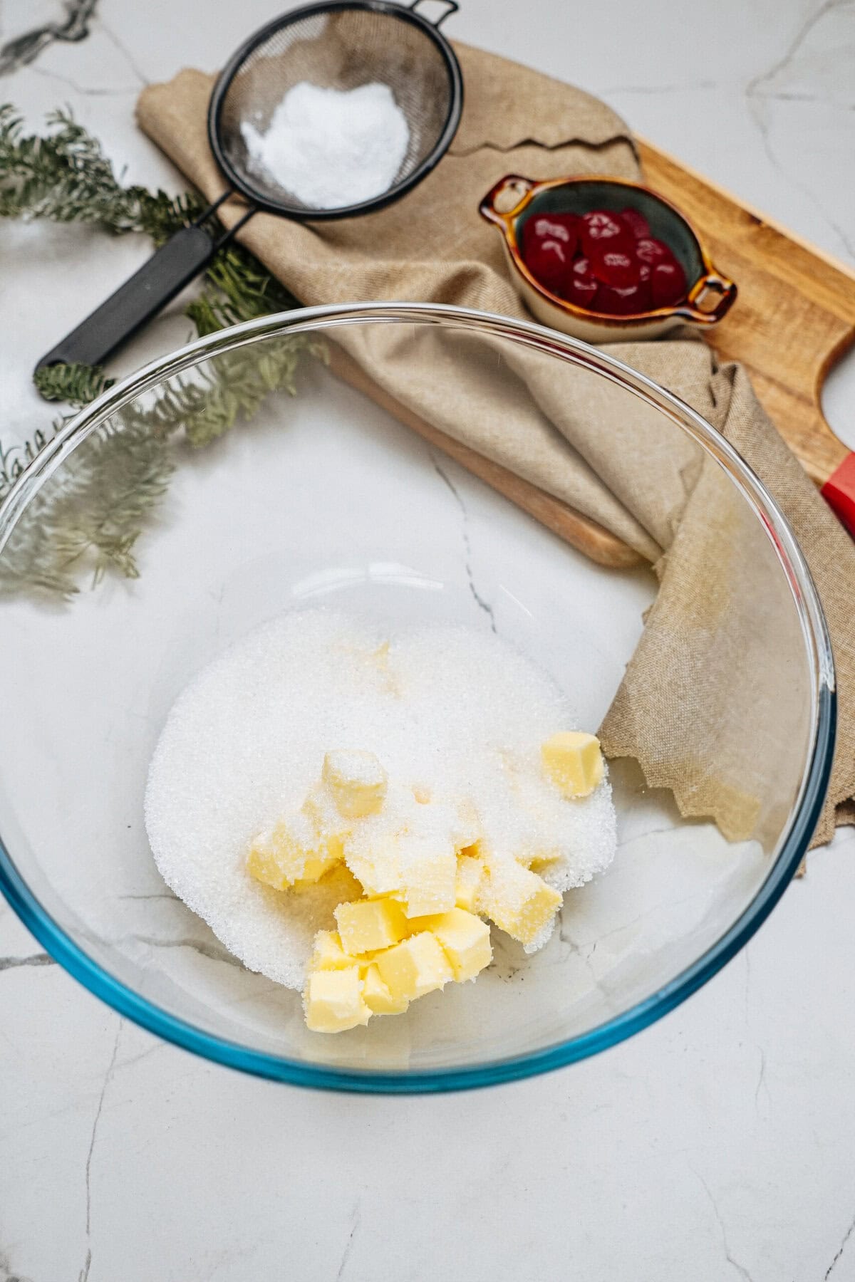 A glass bowl with sugar and butter on a marble surface. Nearby, a sieve with flour, a small dish with red sauce, greenery, and a brown napkin are visible.