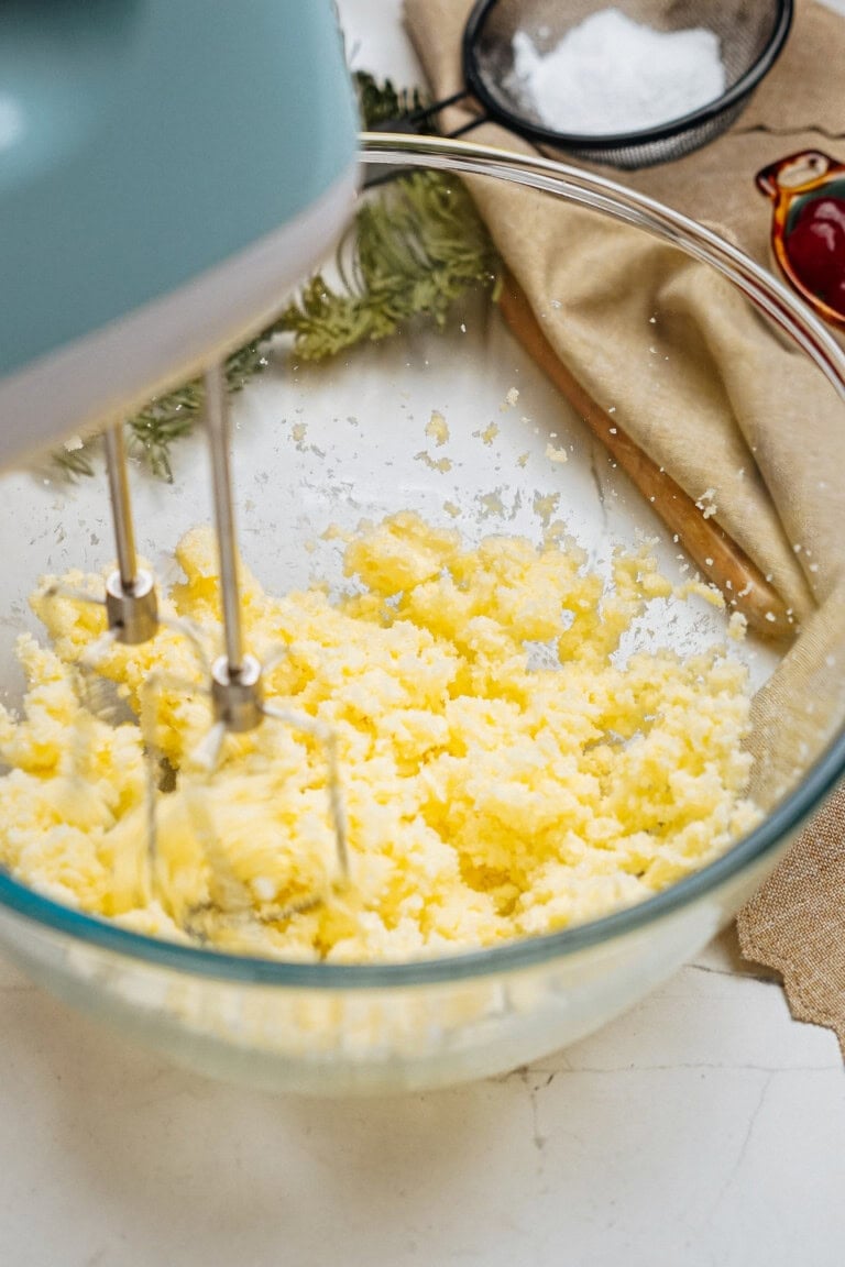 A hand mixer blends butter and sugar in a glass bowl, set on a kitchen countertop with a beige cloth and a small sieve nearby.